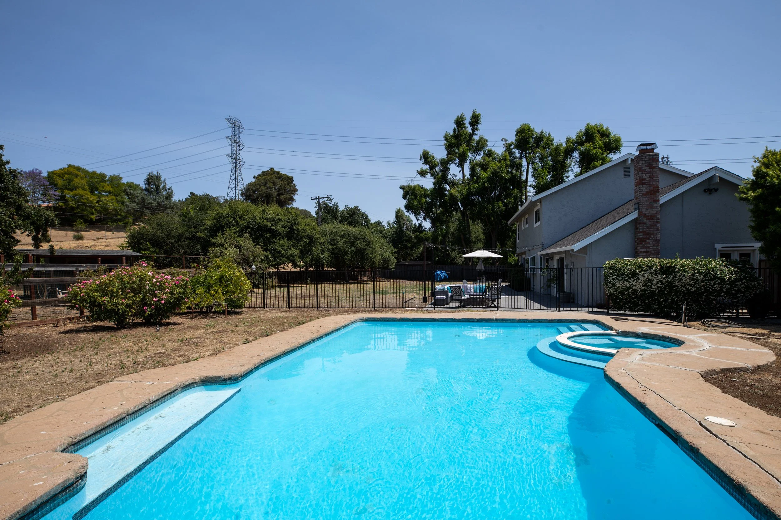 Backyard with swimming pool, patio furniture under a white umbrella, house with chimney, and trees under a clear blue sky.