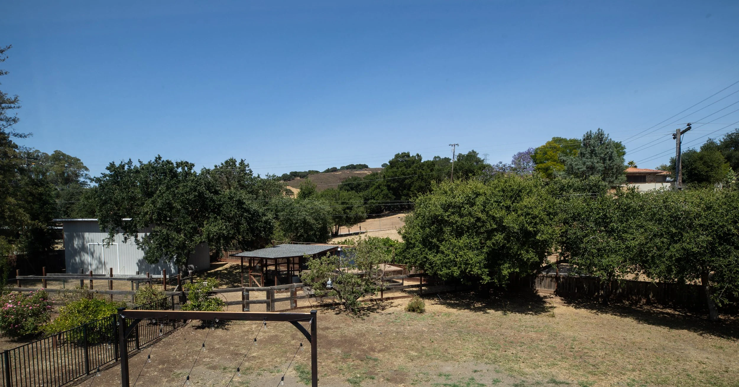 A backyard with trees, a fenced garden area, a shed, and a small structure with a roof. Hills are visible in the background under a clear blue sky.