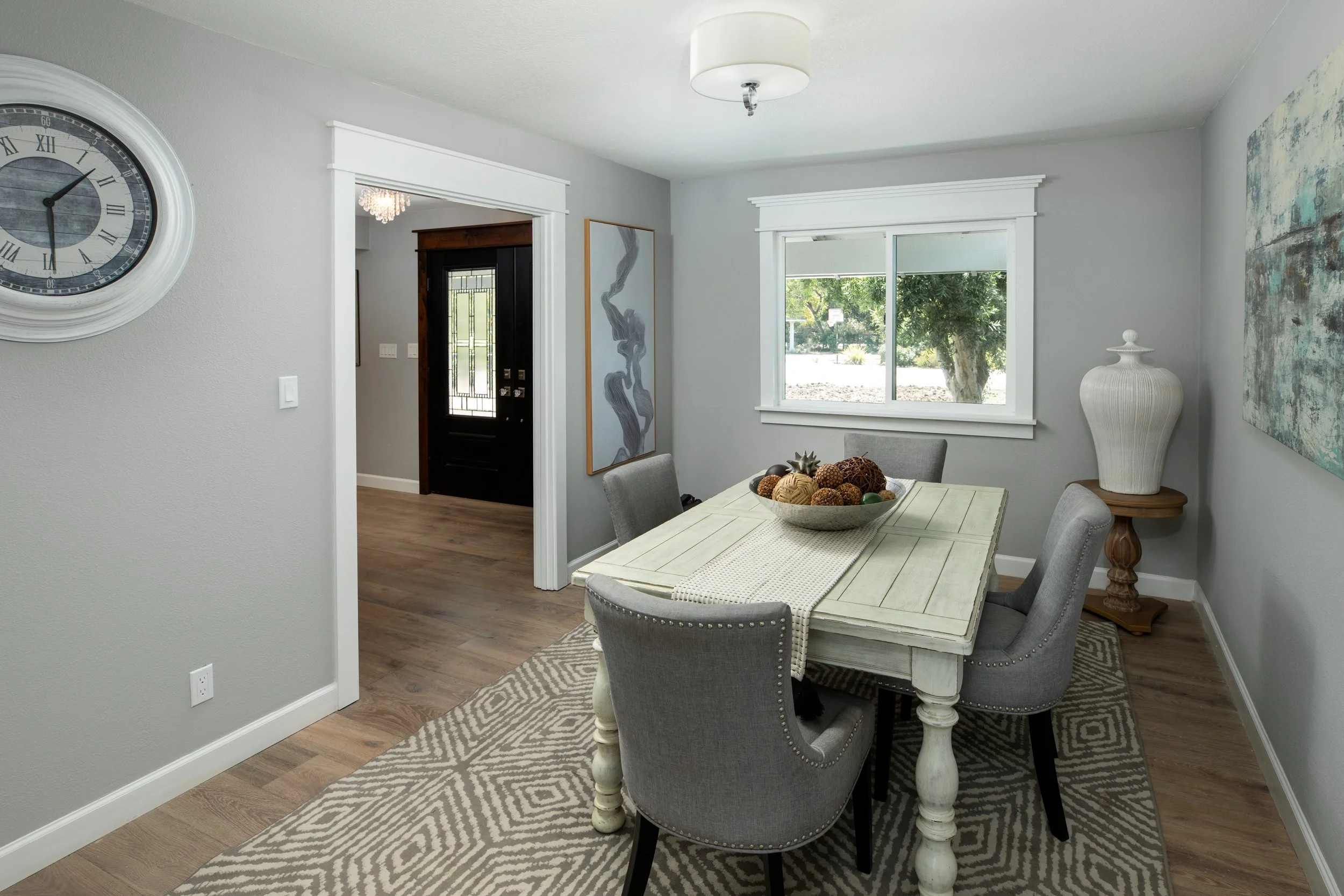 Dining room with a white wooden table, four gray upholstered chairs, a patterned rug, a large bowl of decorative balls on the table, a window with a view of a yard, a white ceramic vase on a small wooden table, artwork on the walls, and a door leadin