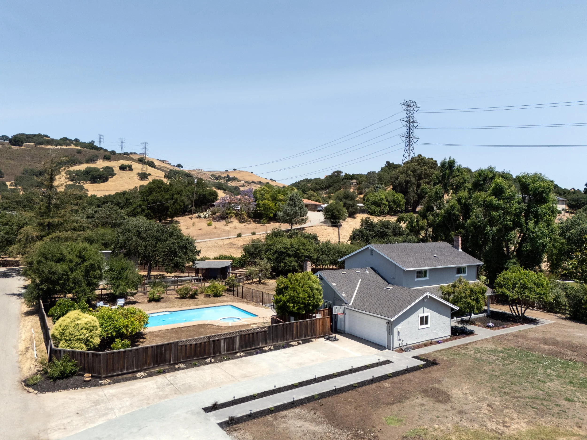 A suburban backyard with a swimming pool, surrounded by a wooden privacy fence, near a two-story gray house with a basketball hoop, with hills and power lines in the background.