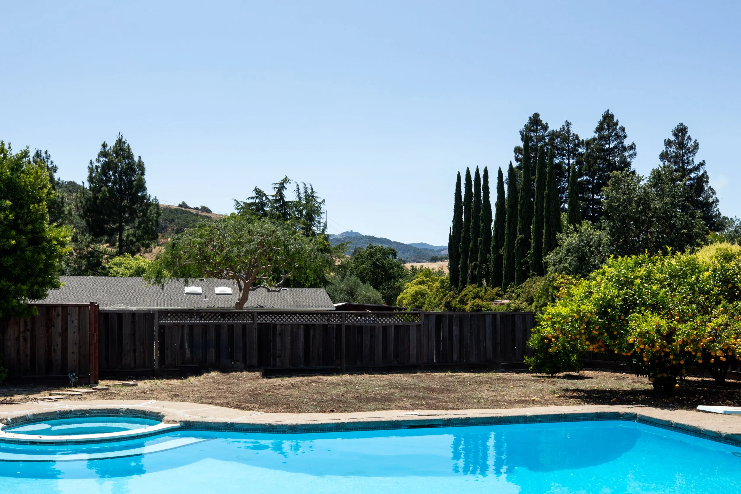 Backyard with a swimming pool, trees, and a wooden fence under a clear blue sky.
