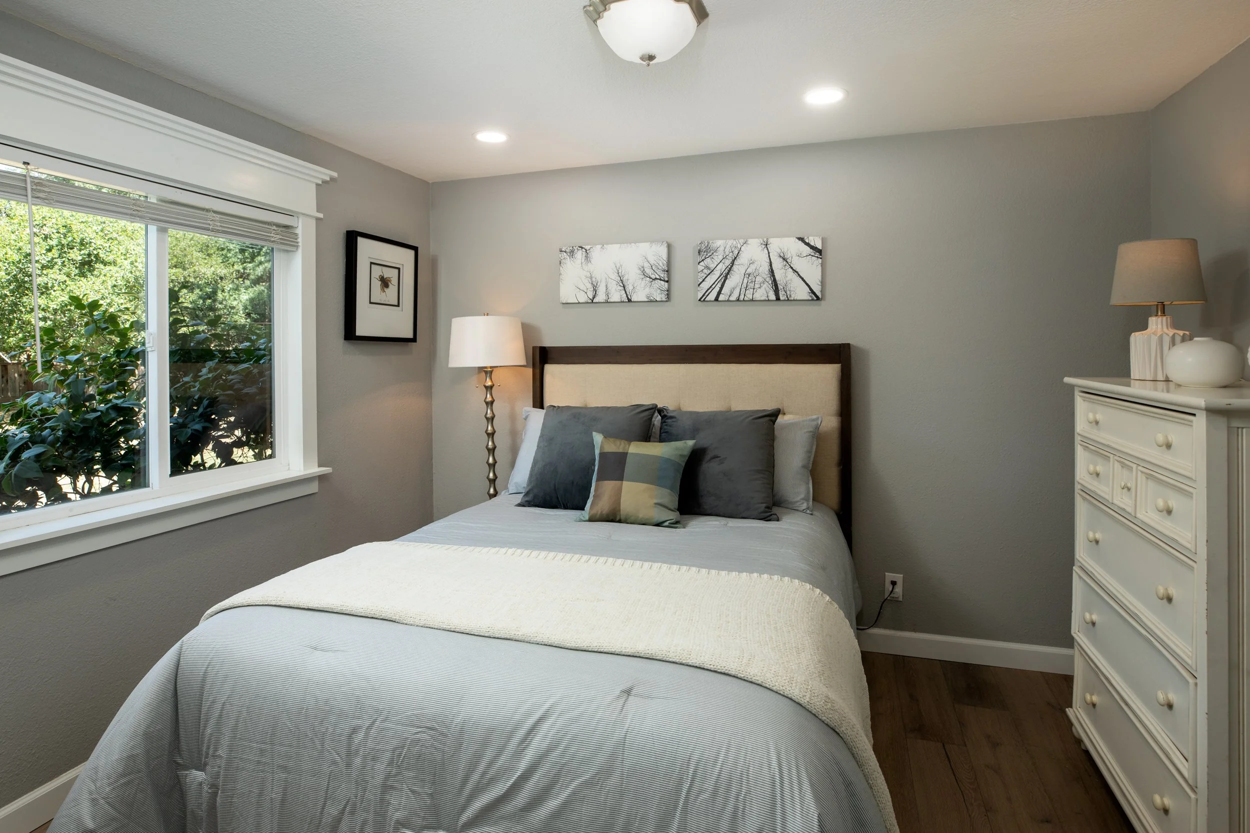 Bedroom with a bed featuring a beige headboard, gray and patterned pillows, a white blanket, and a window with blinds overlooking greenery, with wall art and lamps on side tables and dresser.