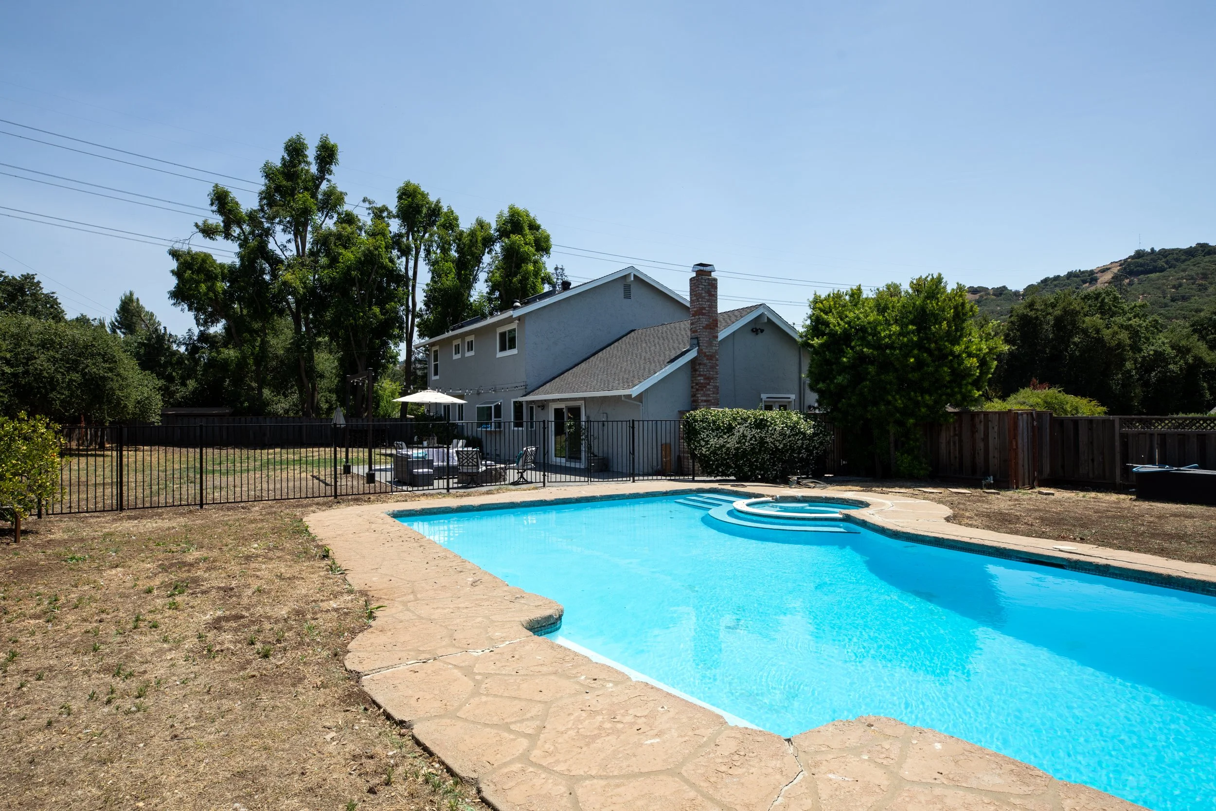 Residential backyard with a blue swimming pool, poolside seating area with umbrella, fenced yard, trees, and a gray house with a chimney in the background under a clear sky.