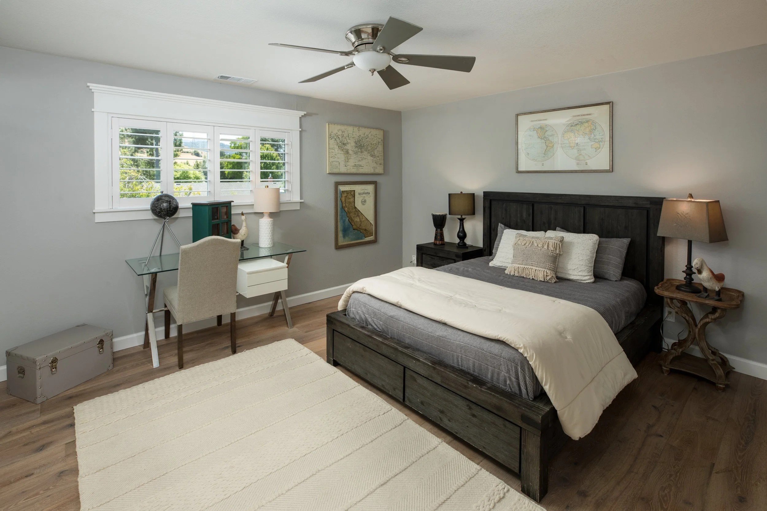 A cozy bedroom with a dark wood bed, beige and gray bedding, and matching nightstands with lamps. The room has light gray walls, a ceiling fan, a window with white shutters, framed world maps, a glass desk with a beige chair, and a beige rug on wood 
