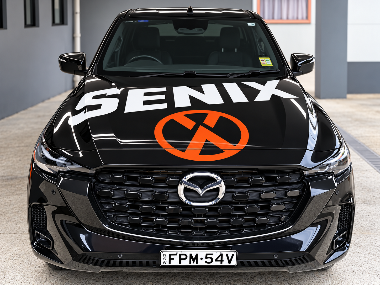 Black Mazda car with custom decal on hood displaying the word 'SENIX' and an orange circle with a black slash through a crossed-out door. The front license plate reads 'FPM-54V.'