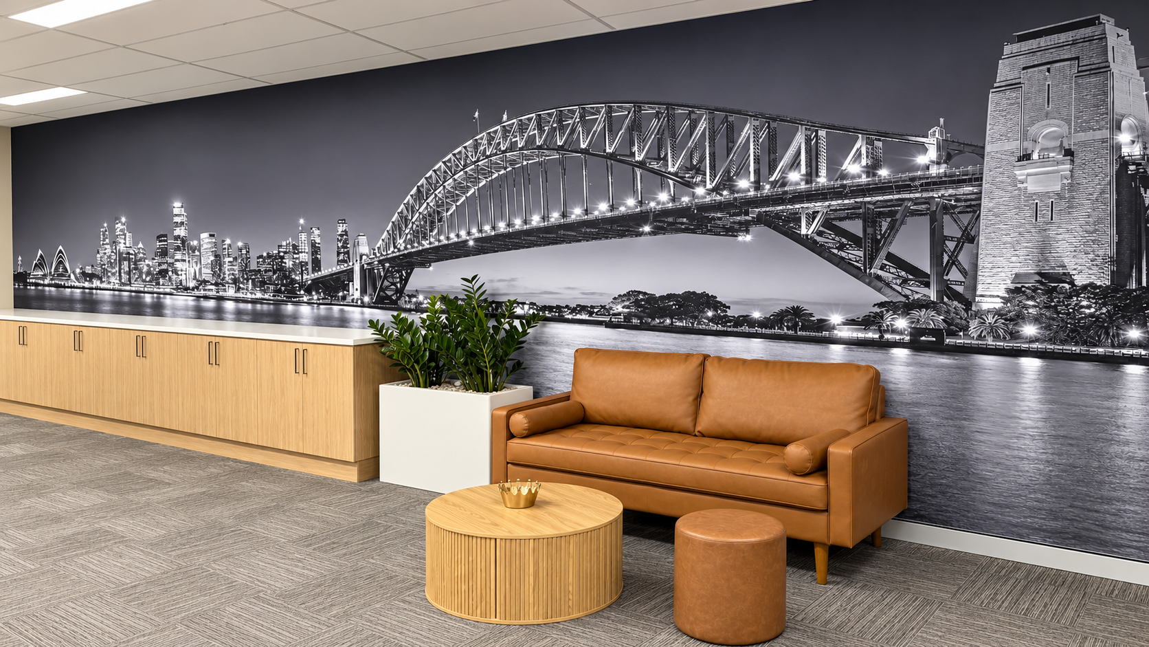 Indoor office lounge area with a large black and white mural of Sydney Harbour Bridge and the city skyline, a brown leather sofa, a round wooden coffee table, a small brown ottoman, potted green plants, and wooden cabinets along the wall.