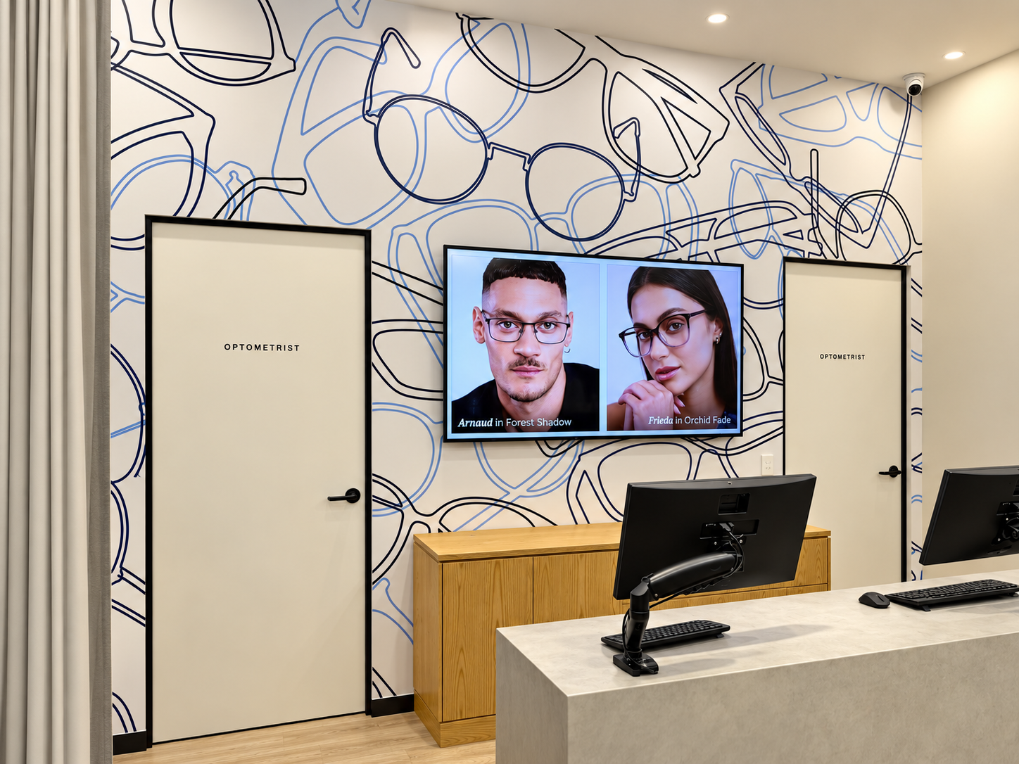 Interior of an optometry office with two closed doors labeled 'Optometrist', a digital display screen showing images of two people wearing glasses with reading text, and desks with computer monitors.