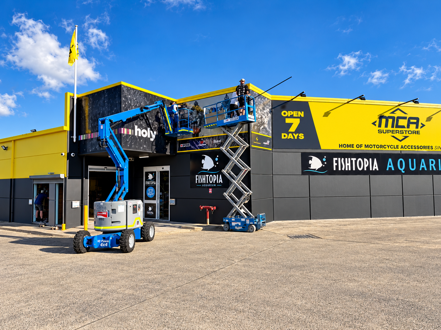People working on the exterior of a store called Fishtopia Aquarium, with banners announcing it's open for 7 days, under a bright blue sky with some clouds.