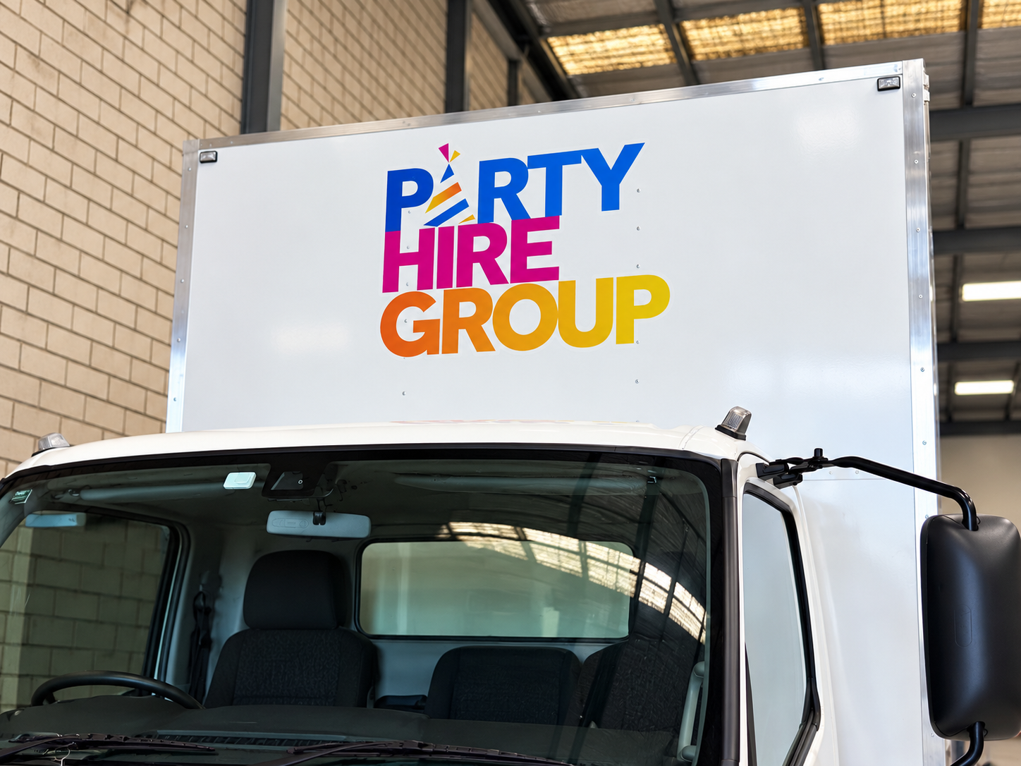 A white truck with a large sign on the top that reads 'Party Hire Group' in colorful letters, parked inside a warehouse or large garage.