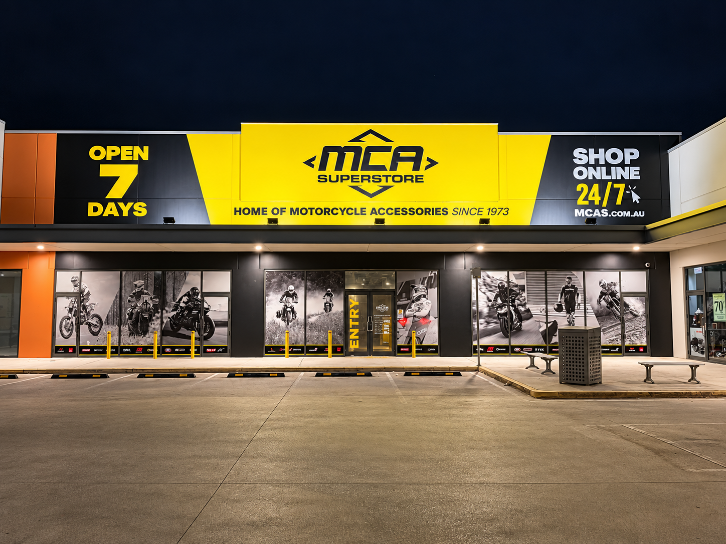 Front view of MCA Superstore at night with signage indicating 7 days open, online shopping 24/7, and images of motorcyclists and accessories on windows.