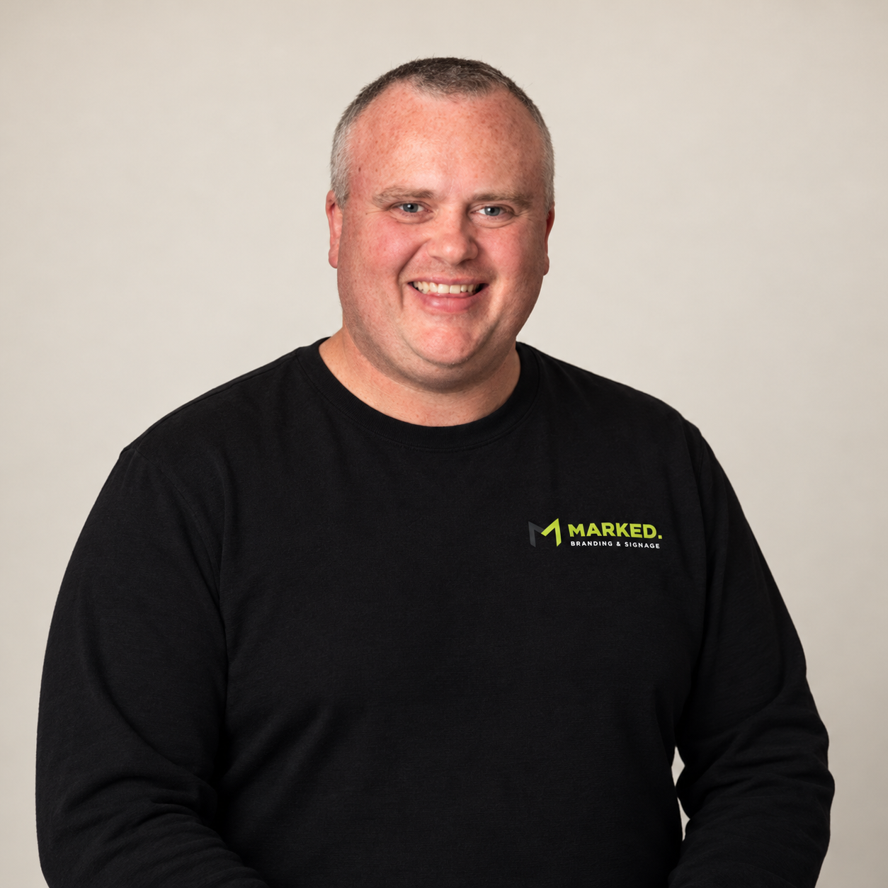 A man with short light hair and a big smile, wearing a black long-sleeve shirt with a logo that says 'MARKED Branding & Signage' on the chest, standing against a plain light gray background.