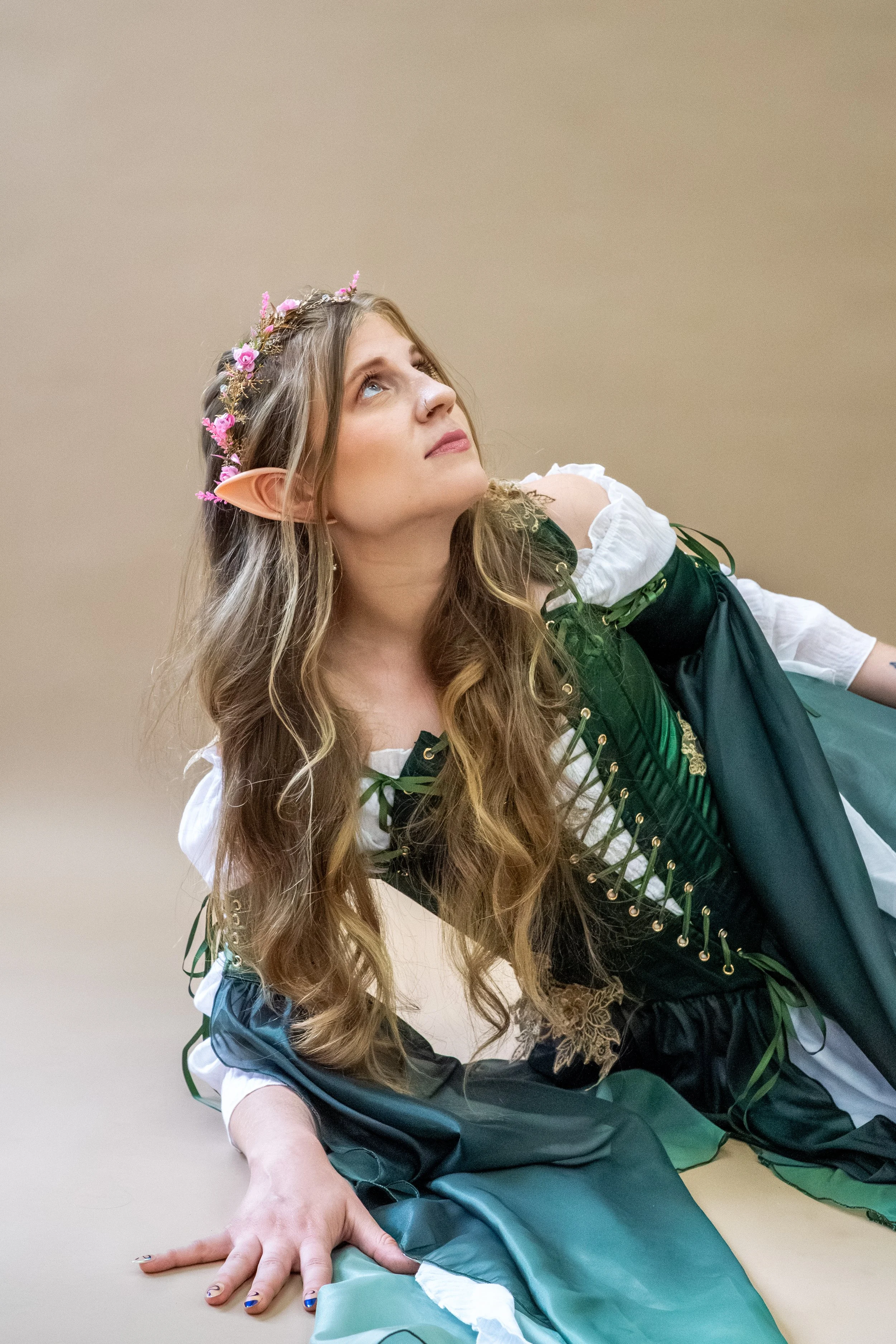Woman wearing a pink floral crown, pointed elf ears, and a green fantasy costume, looking upward