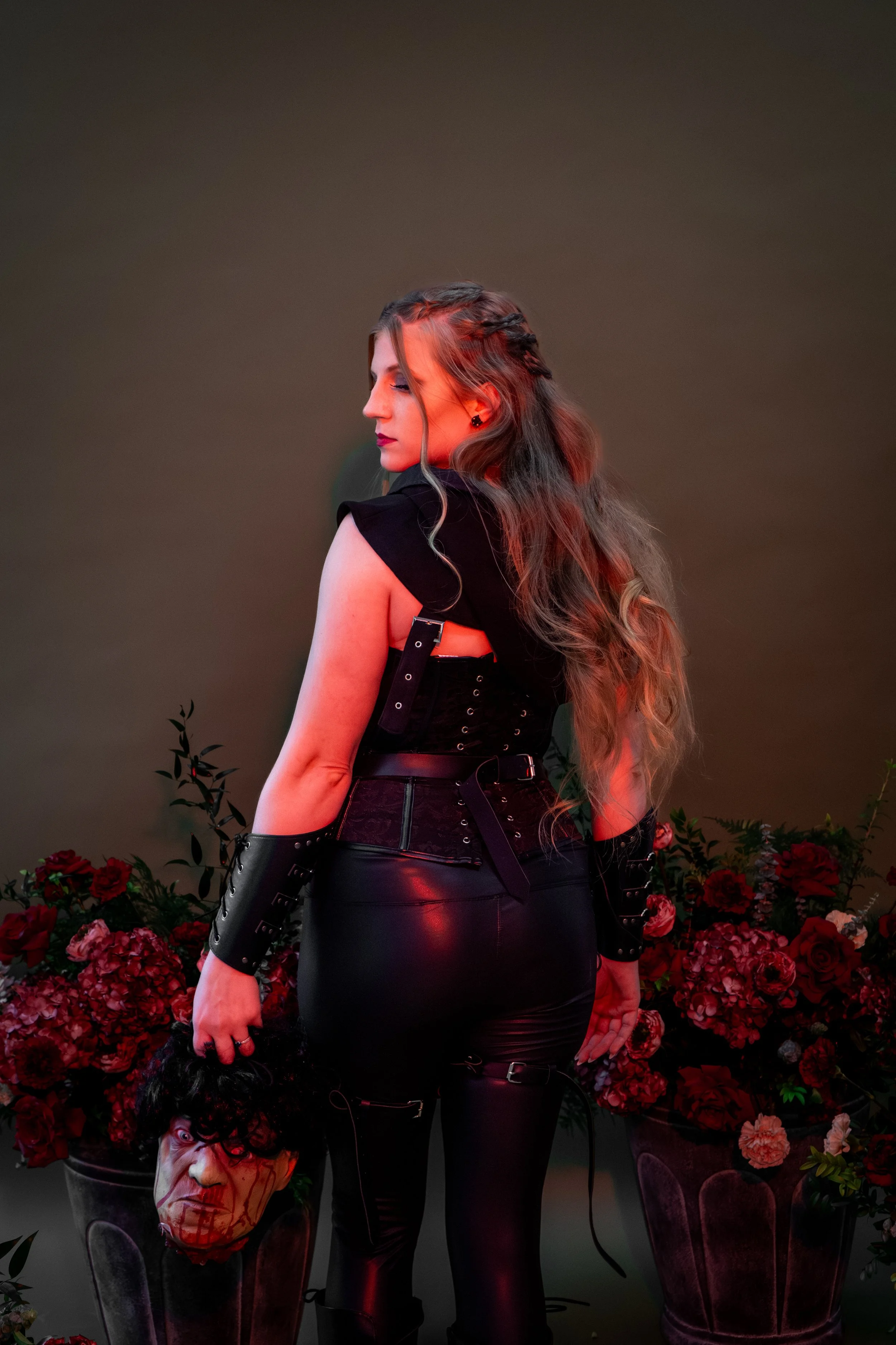 Woman in black gothic outfit with braided hair standing among red flowers, holding a bloodied severed head prop.