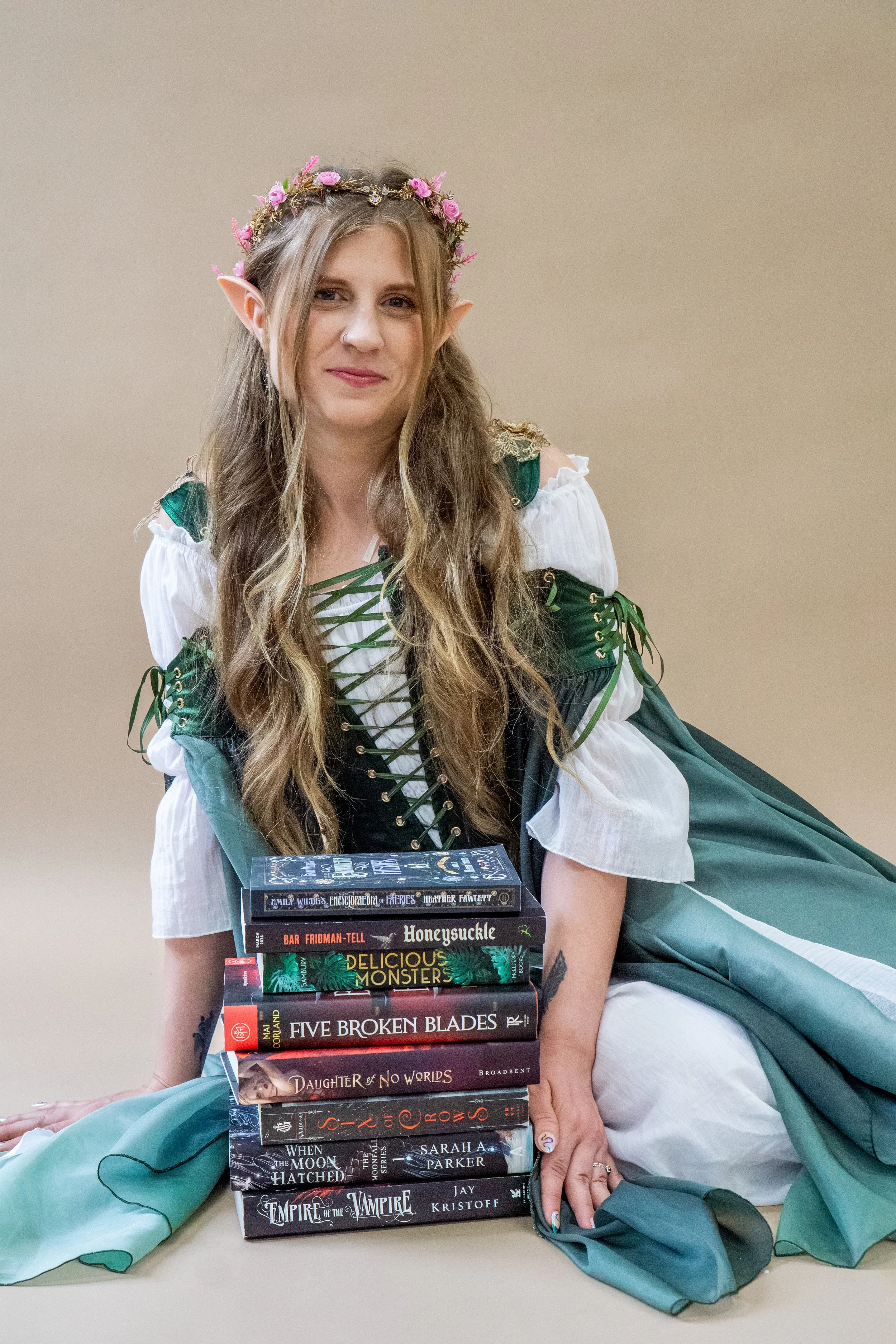 Woman dressed as a flower-crowned elf in a green Renaissance-style costume, sitting behind a stack of fantasy novels.