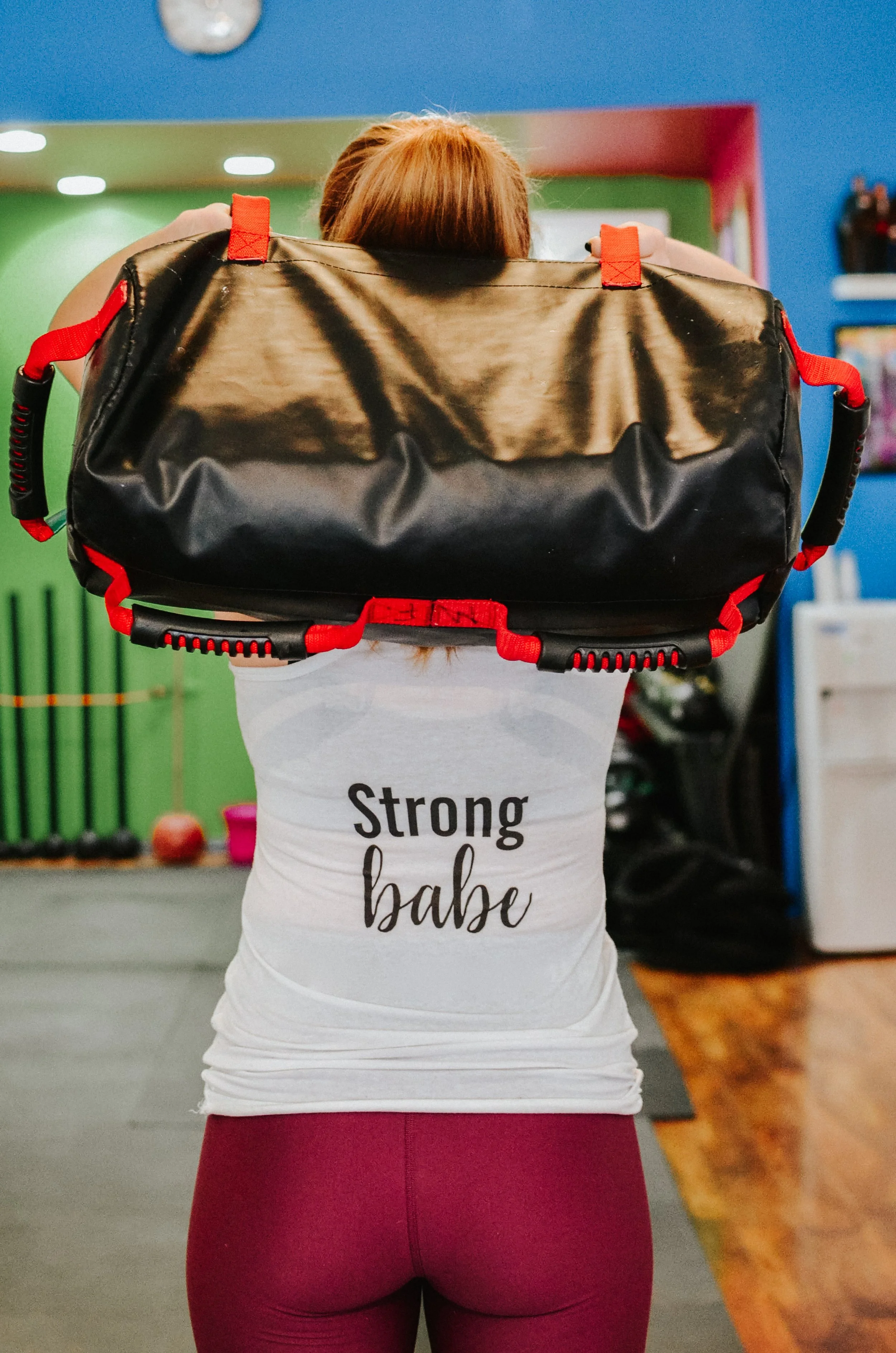 Person seen from behind carrying a large black duffel bag with red straps in a colorful gym.