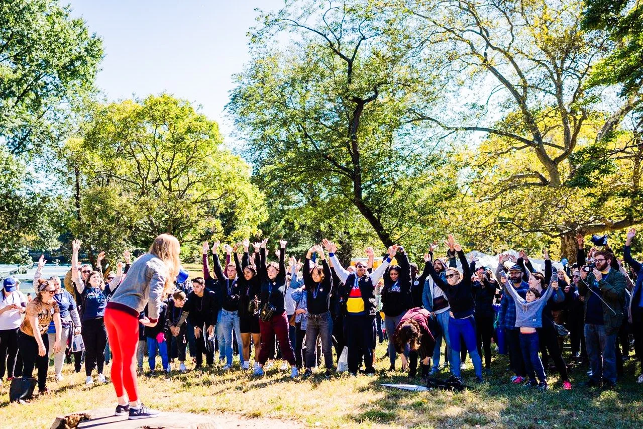 Outdoor group fitness class in a park with participants raising their hands and an instructor leading exercises