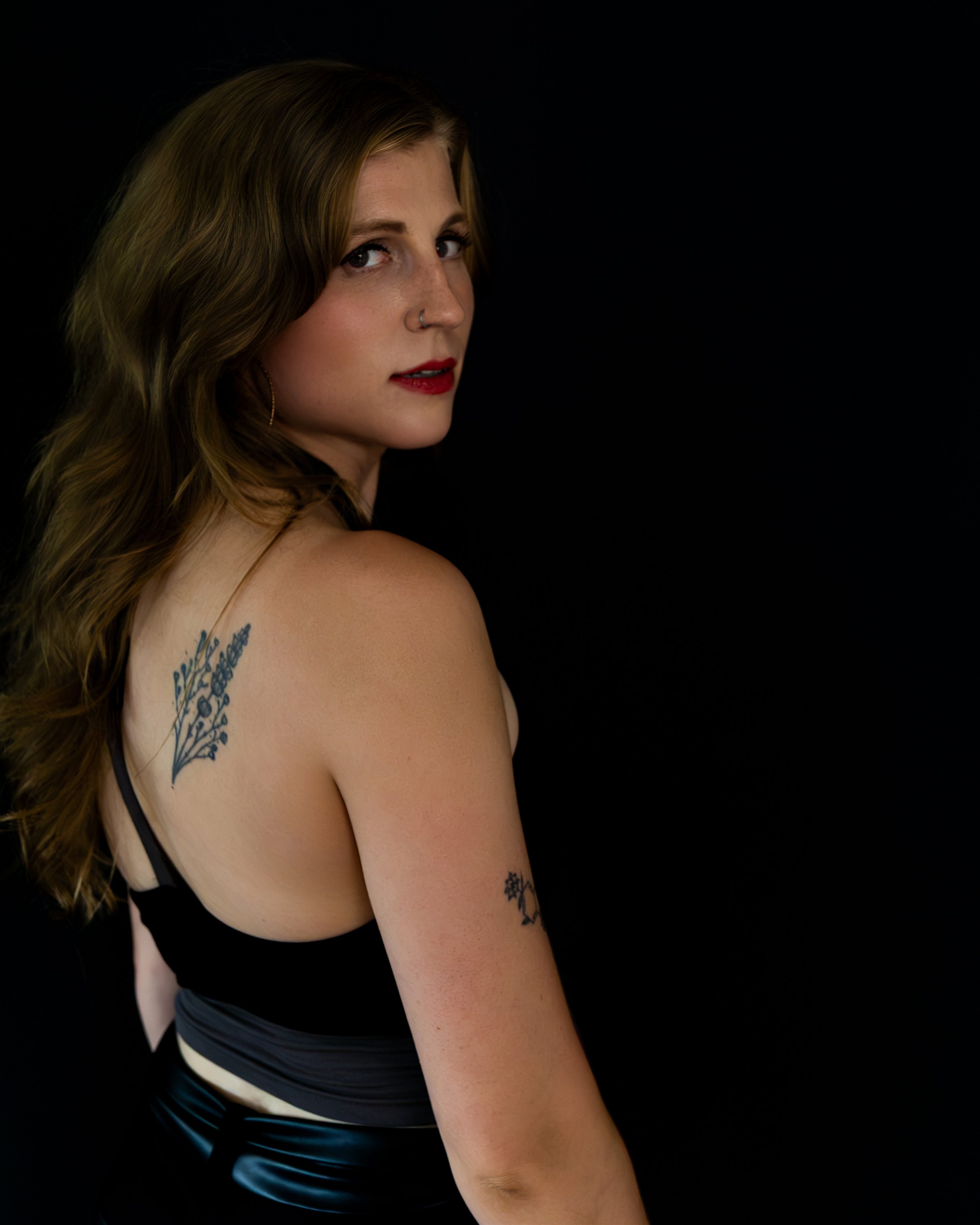 Portrait of a woman with wavy brown hair, red lipstick, nose ring, and shoulder tattoos against a black background