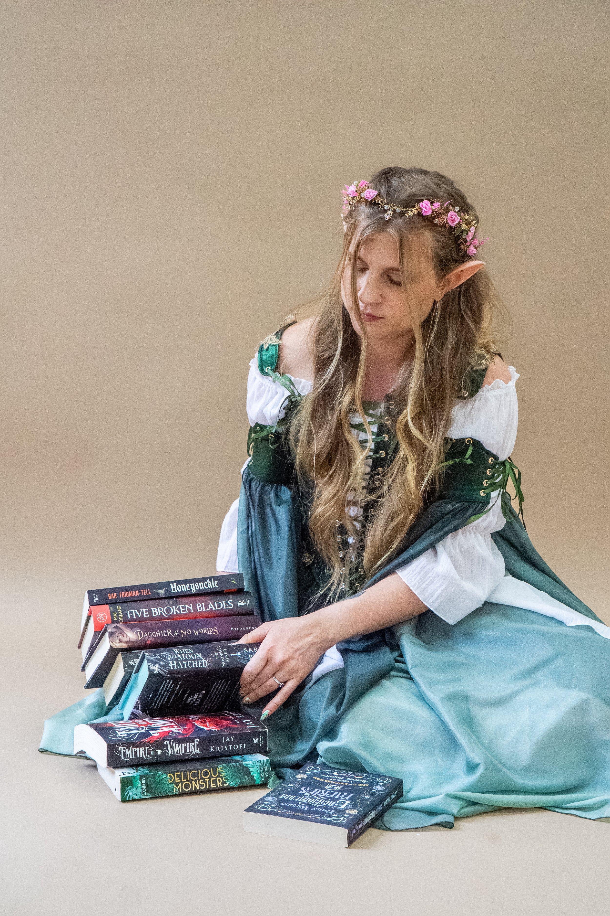 Woman in a green medieval-style dress with pointed elf ears and a pink floral crown, holding a stack of fantasy novels.
