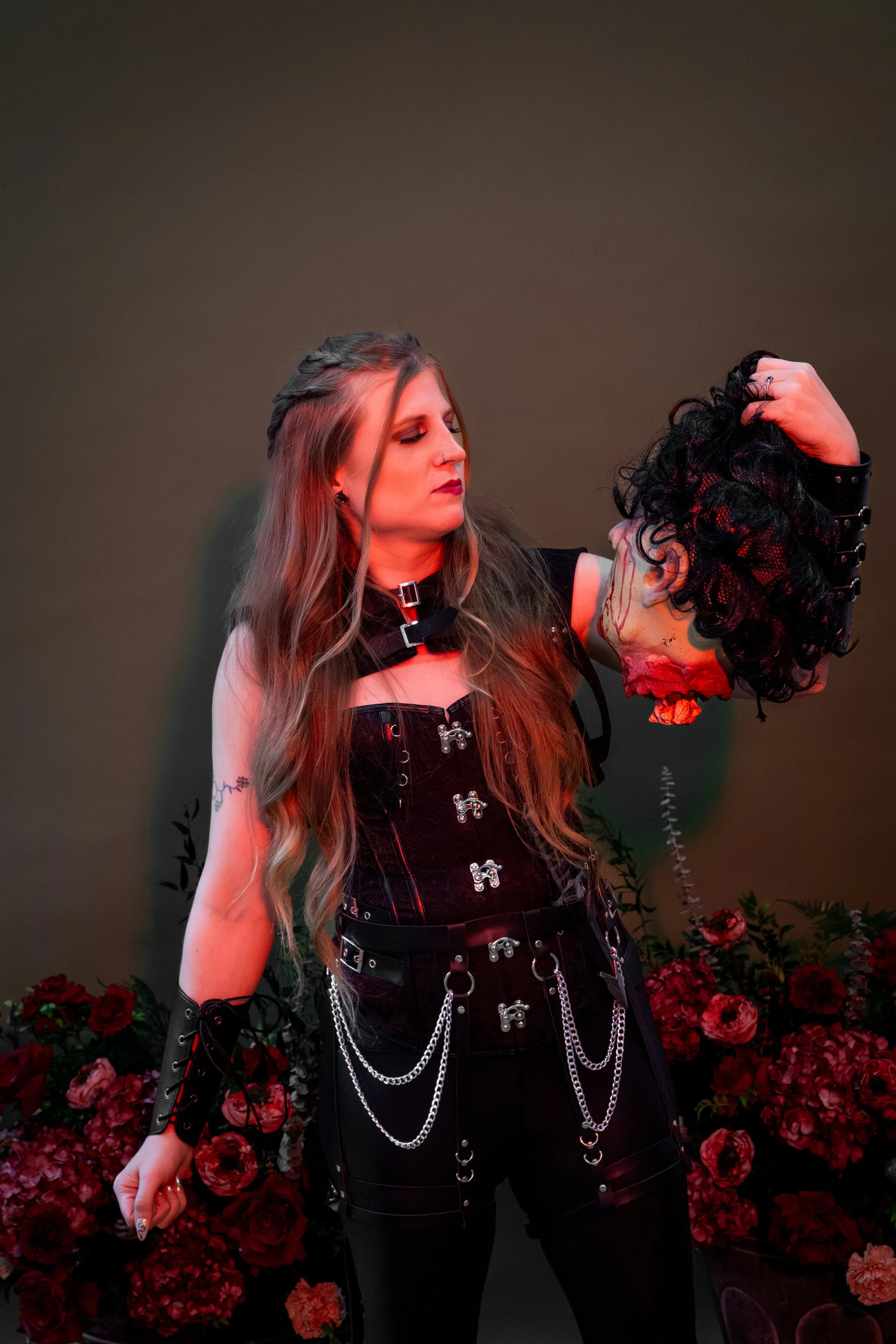 Gothic woman in a black corset and chain accessories holding a severed zombie head prop amid dark red flowers.