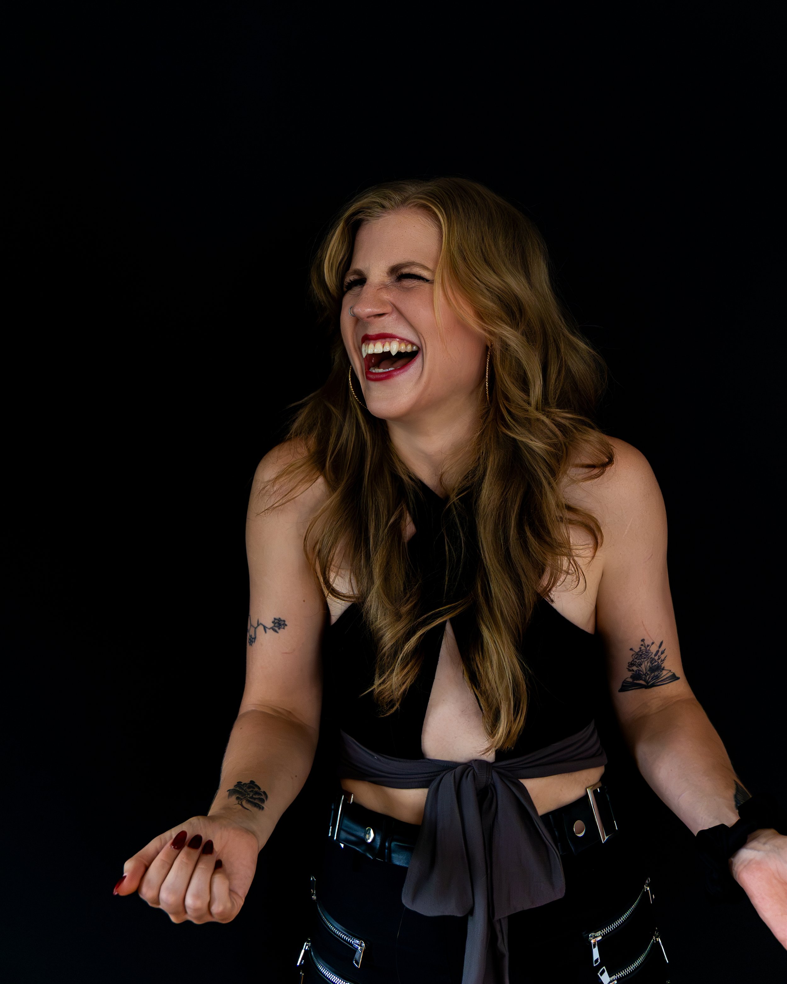 Smiling tattooed woman with long wavy hair and a nose ring laughing against a black background