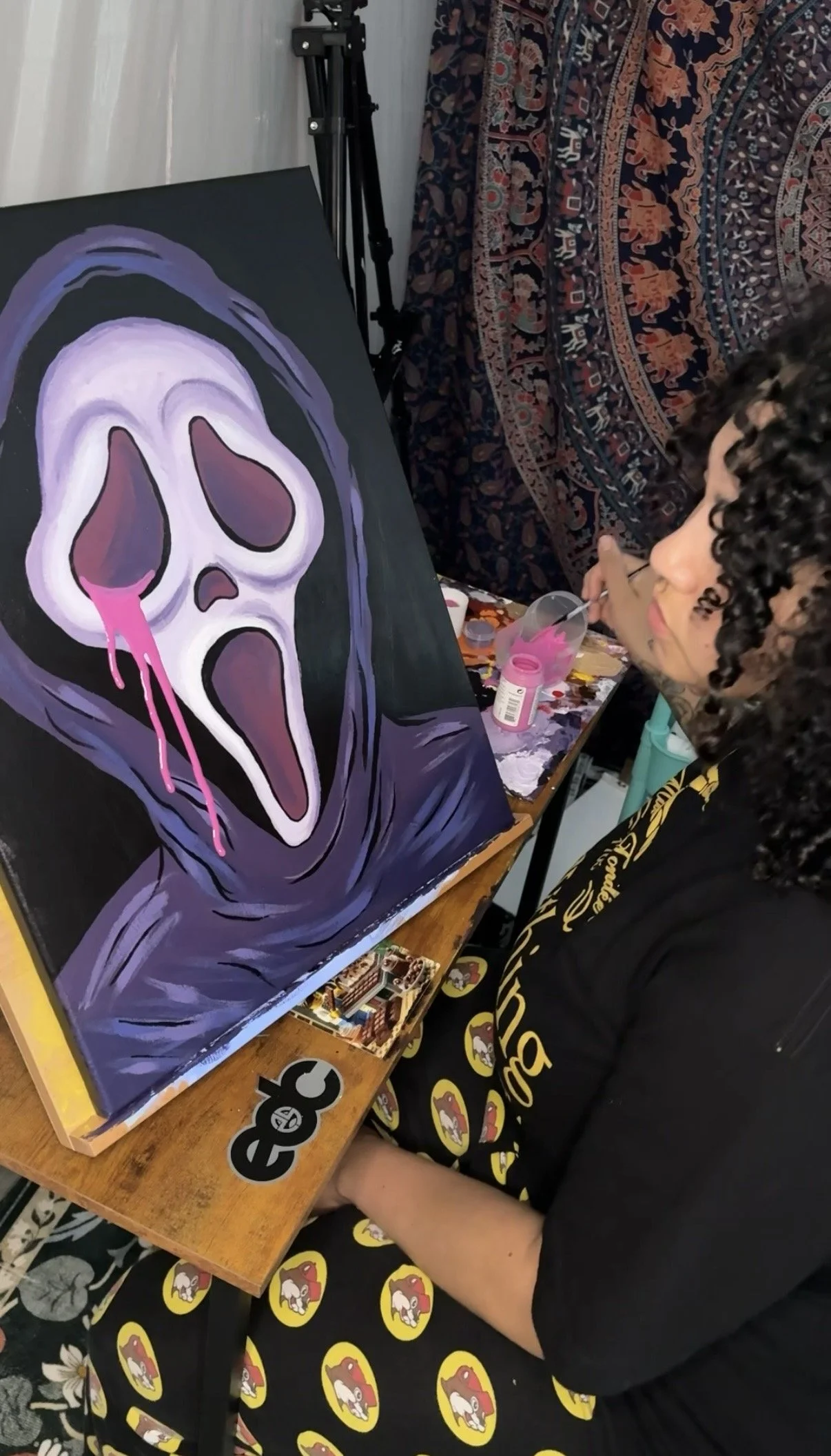 A person with curly hair painting a large canvas of a ghost face with purple and pink tones. The person is sitting at a wooden table covered with stickers and art supplies in a room with a patterned tapestry and a camera tripod.