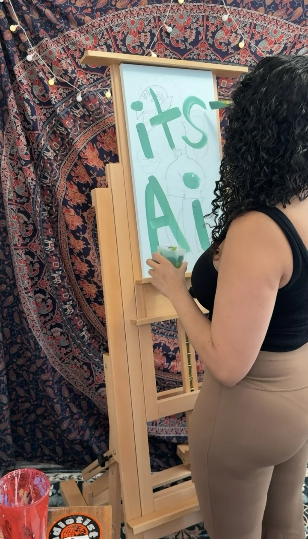 A woman with curly black hair, wearing a black sleeveless top and beige fitted pants, is painting on a whiteboard with large green letters spelling 'Its' and 'Ai'. The background features a colorful, patterned tapestry with elephants and intricate designs, and a string of small decorative lights. A red drink is on the table in front of her.