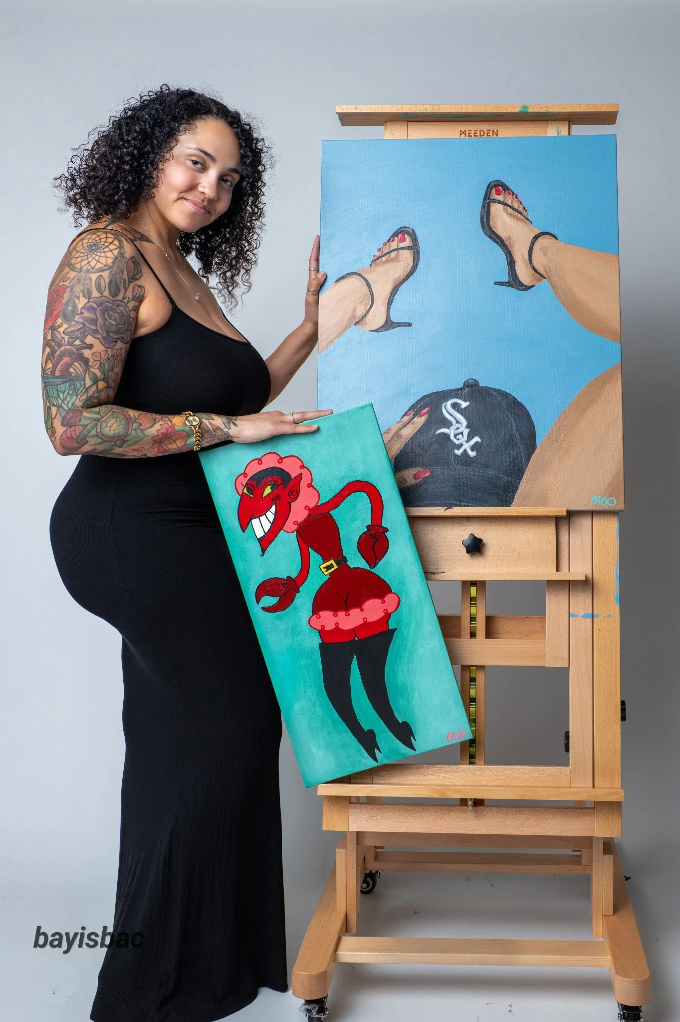 A woman with curly hair, tattoos on her arm, wearing a black dress, is standing next to an easel holding a completed painting of a pair of feet with red nail polish and black high heels, viewed from above, against a blue background. She is holding a painting of a red cartoon devil character with black horns.