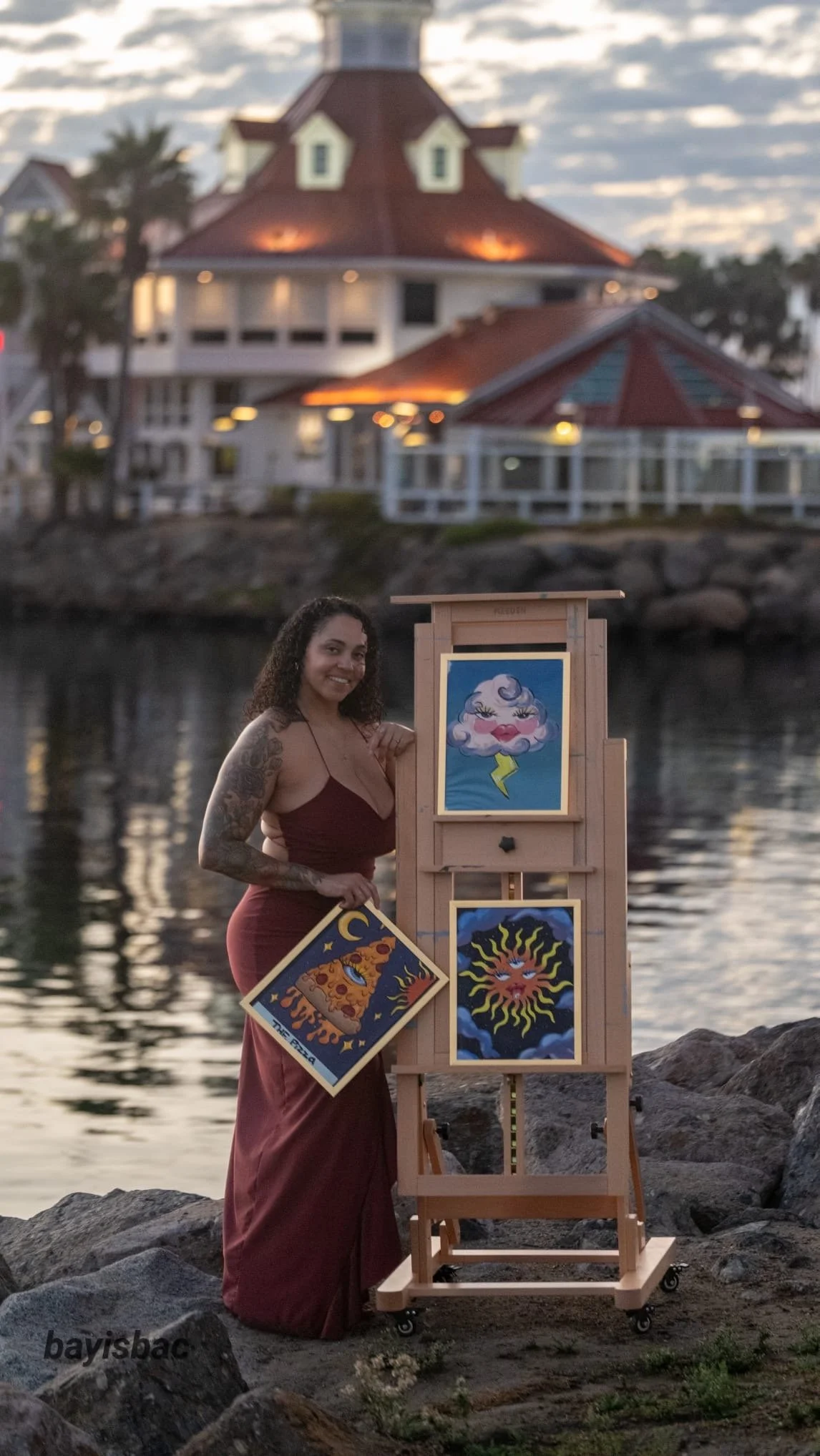 A woman with curly hair and tattoos on her arm standing by an easel with colorful tarot card art, near a body of water and a large house with a red roof in the background during sunset.