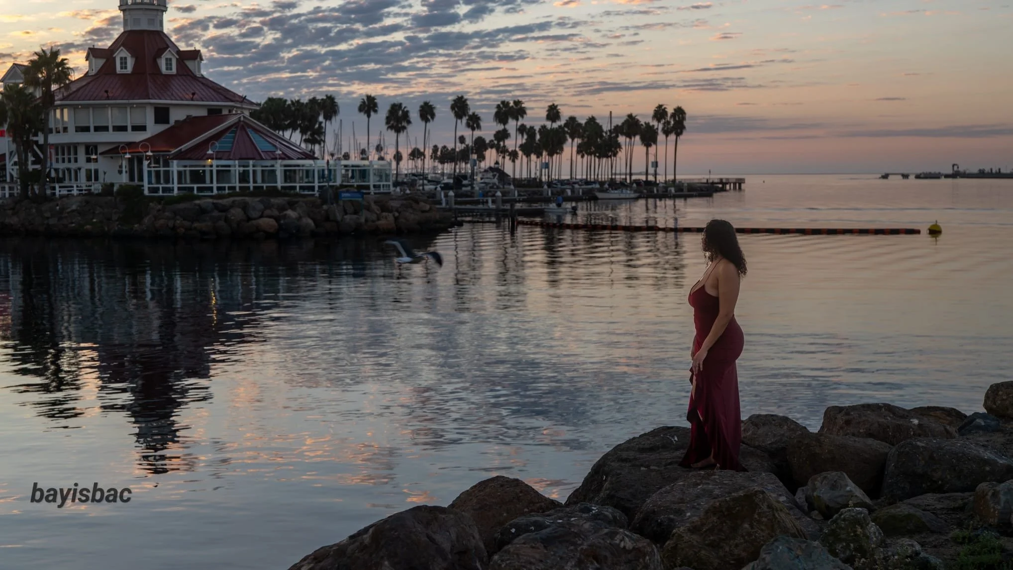A woman in a long red dress standing on rocks by the water at sunset, with palm trees, a building, and boats in the background.