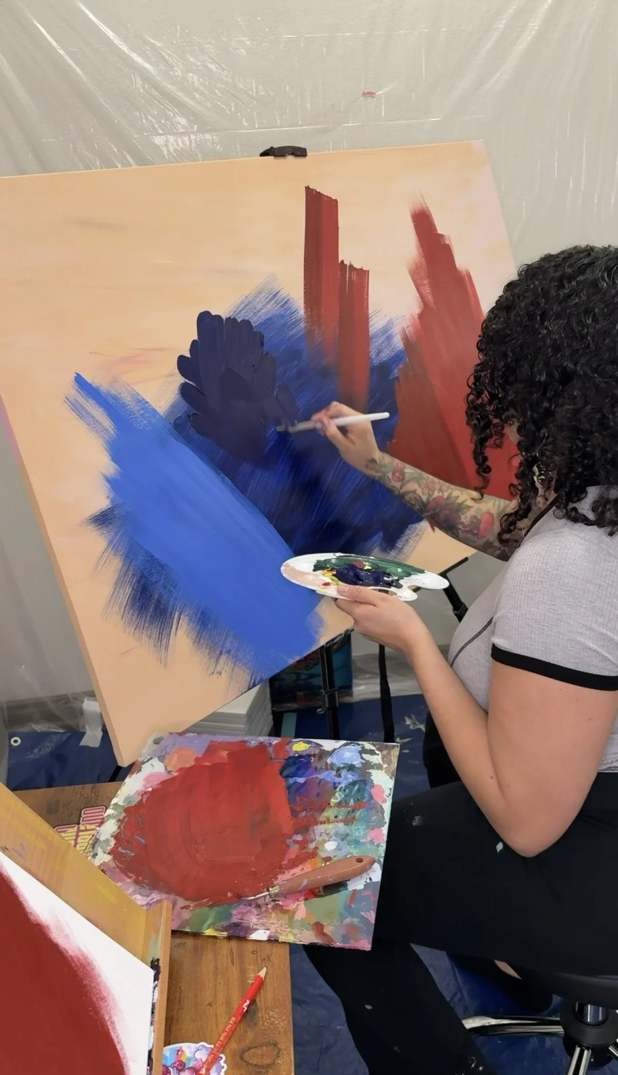 A person with curly hair and tattoos on their arm is painting on a large canvas, using red and blue paints. The workspace includes paint palettes, brushes, and additional paintings, with a plastic sheet covering the wall in the background.