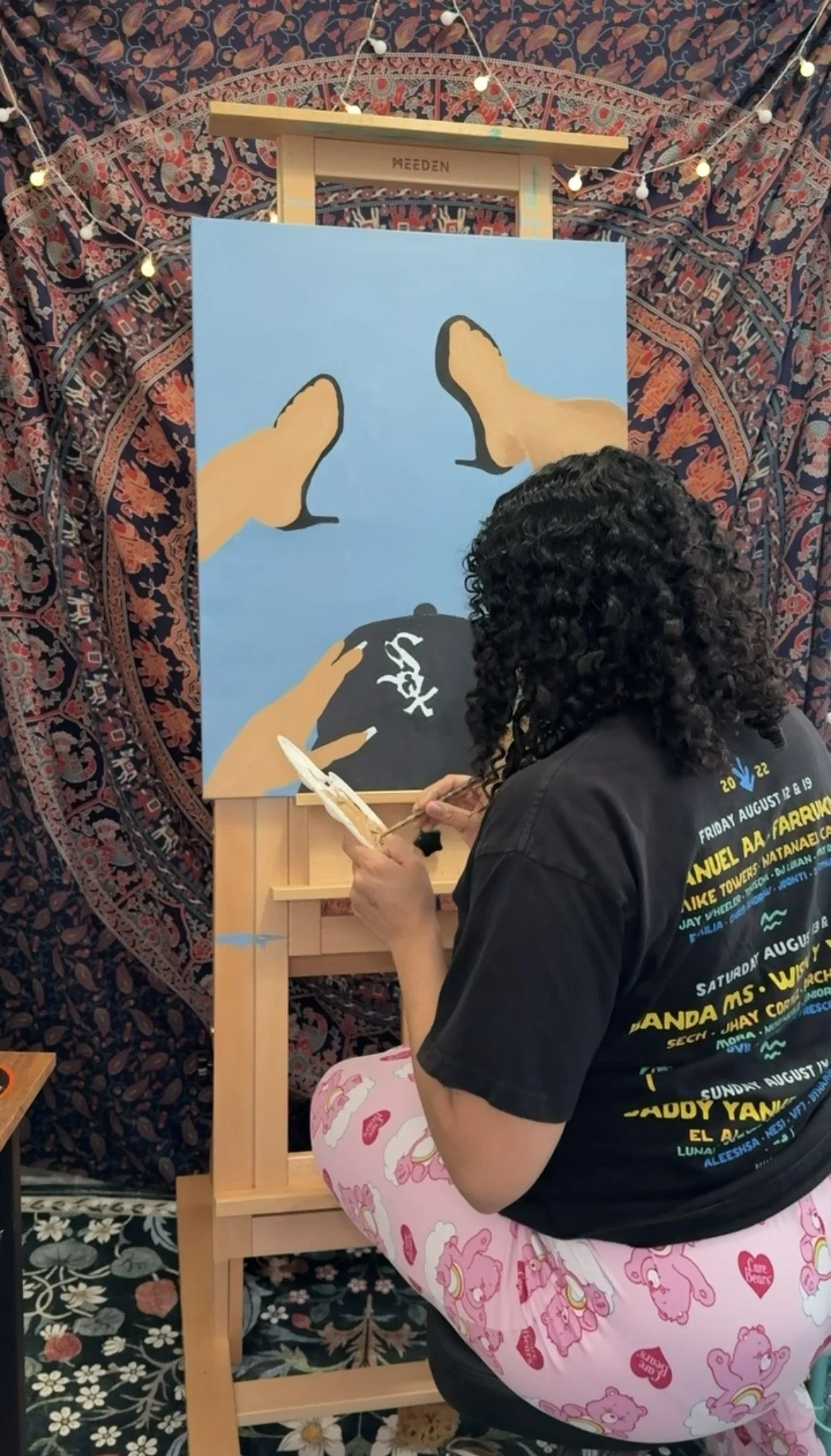 A person with curly hair painting on a canvas with a small brush. The person is sitting on a chair covered with pink fabric featuring various cartoon characters. The background features a colorful, patterned tapestry and string lights hanging overhead.