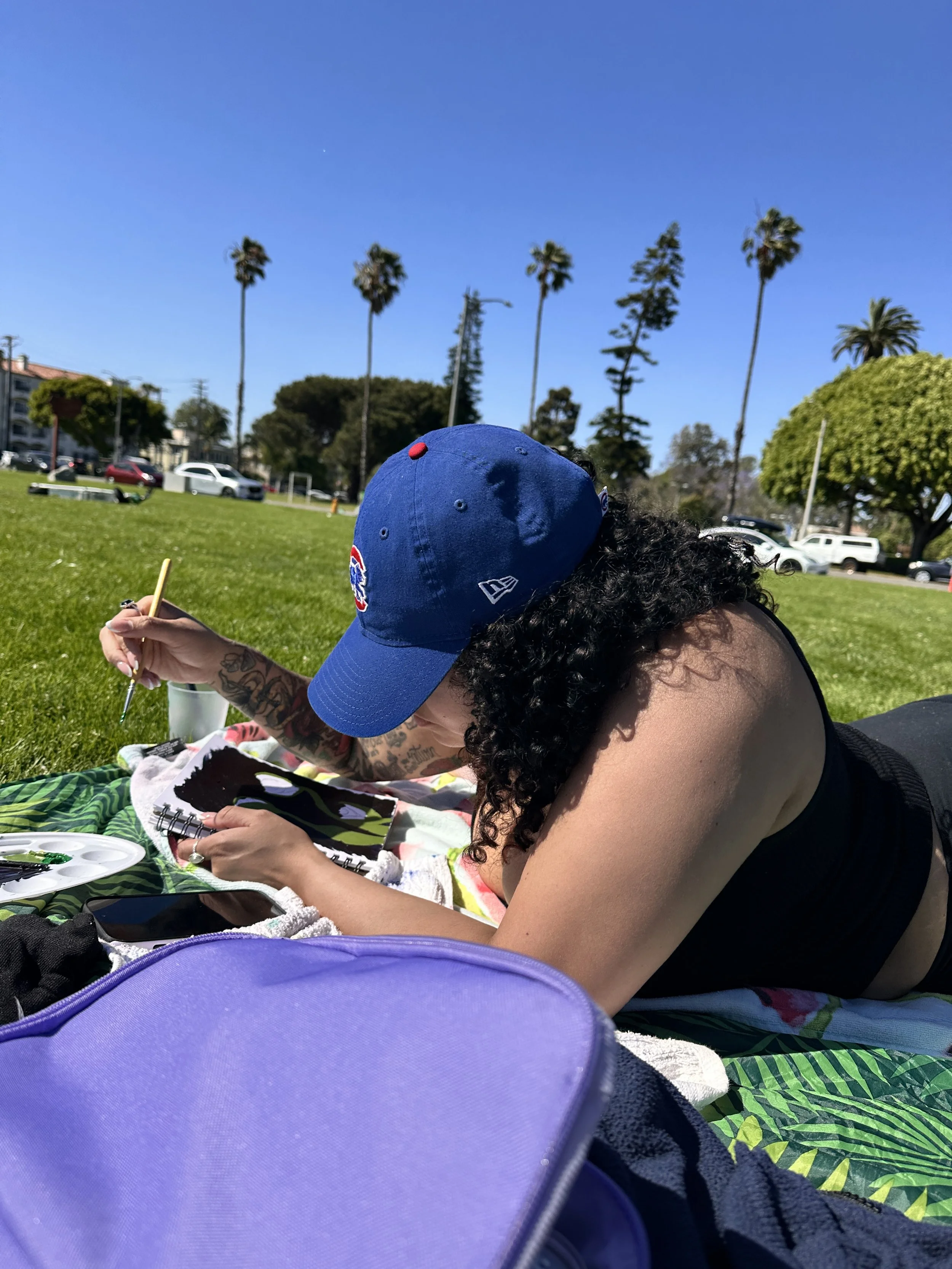 Person with curly hair wearing a blue cap, lying on a colorful towel at a park, painting or drawing on a small sketchbook with a paintbrush, surrounded by various bags and personal items, with green grass, palm trees, and parked cars in the background under a clear blue sky.