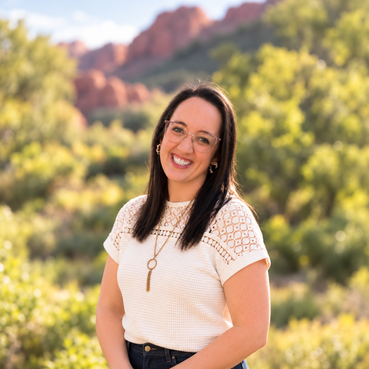 A woman with dark hair, glasses, and a white lace top, smiling outdoors with trees and mountains in the background.