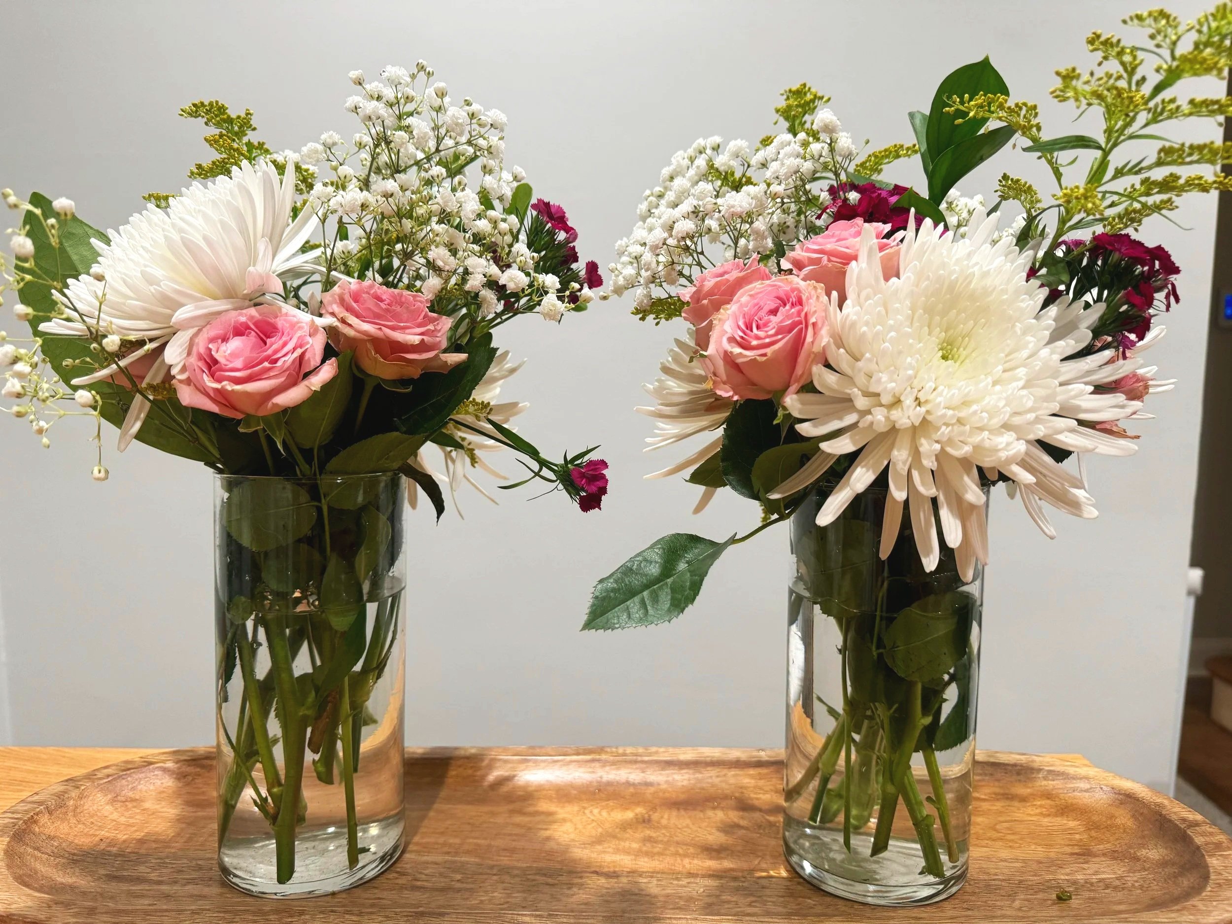 Three glass vases with floral arrangements on a wooden board, with white, pink, and purple flowers and green leaves.