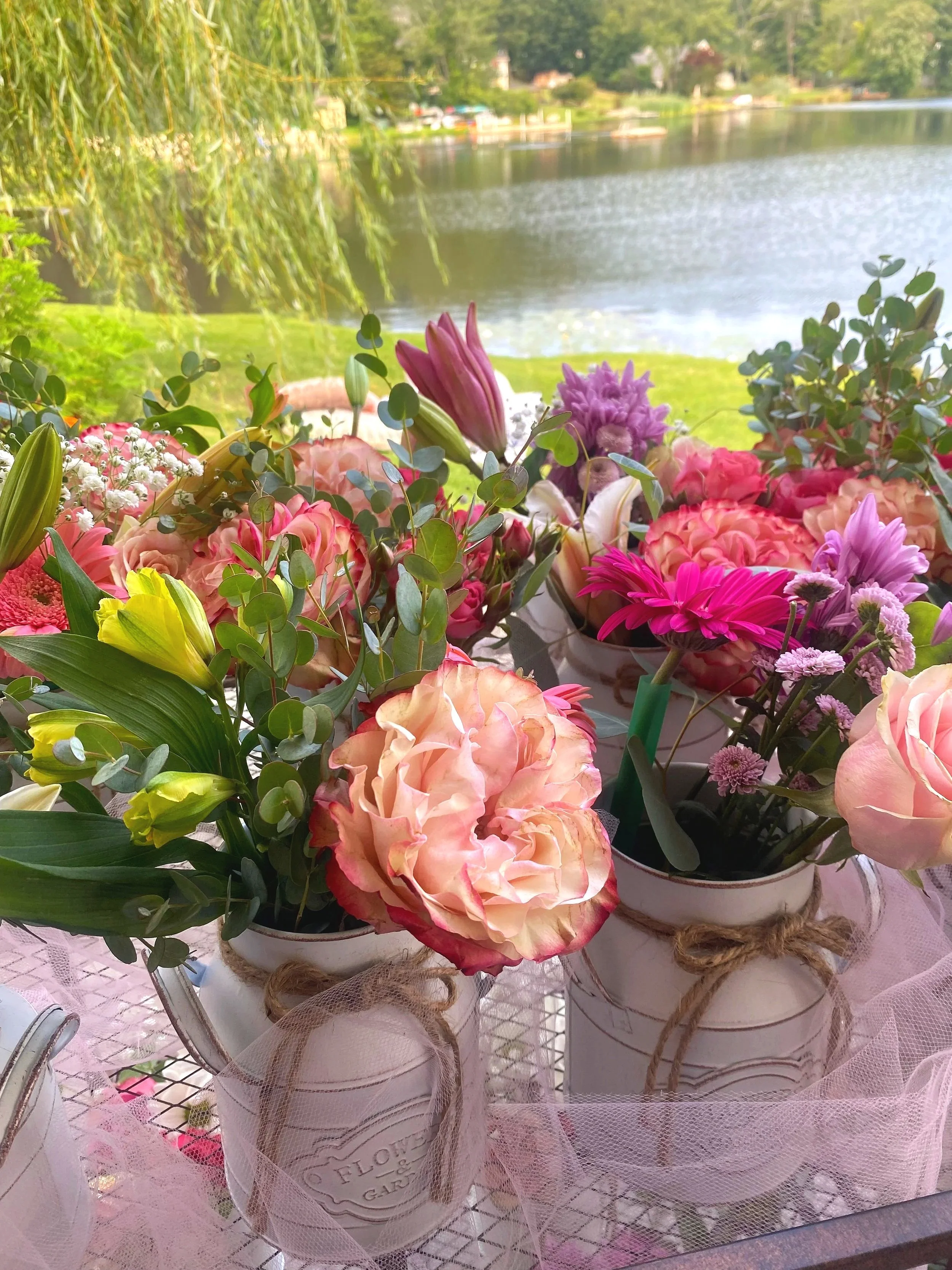 Colorful bouquets of flowers in white vases with twine, placed on a table near a lake with trees and houses in the background.