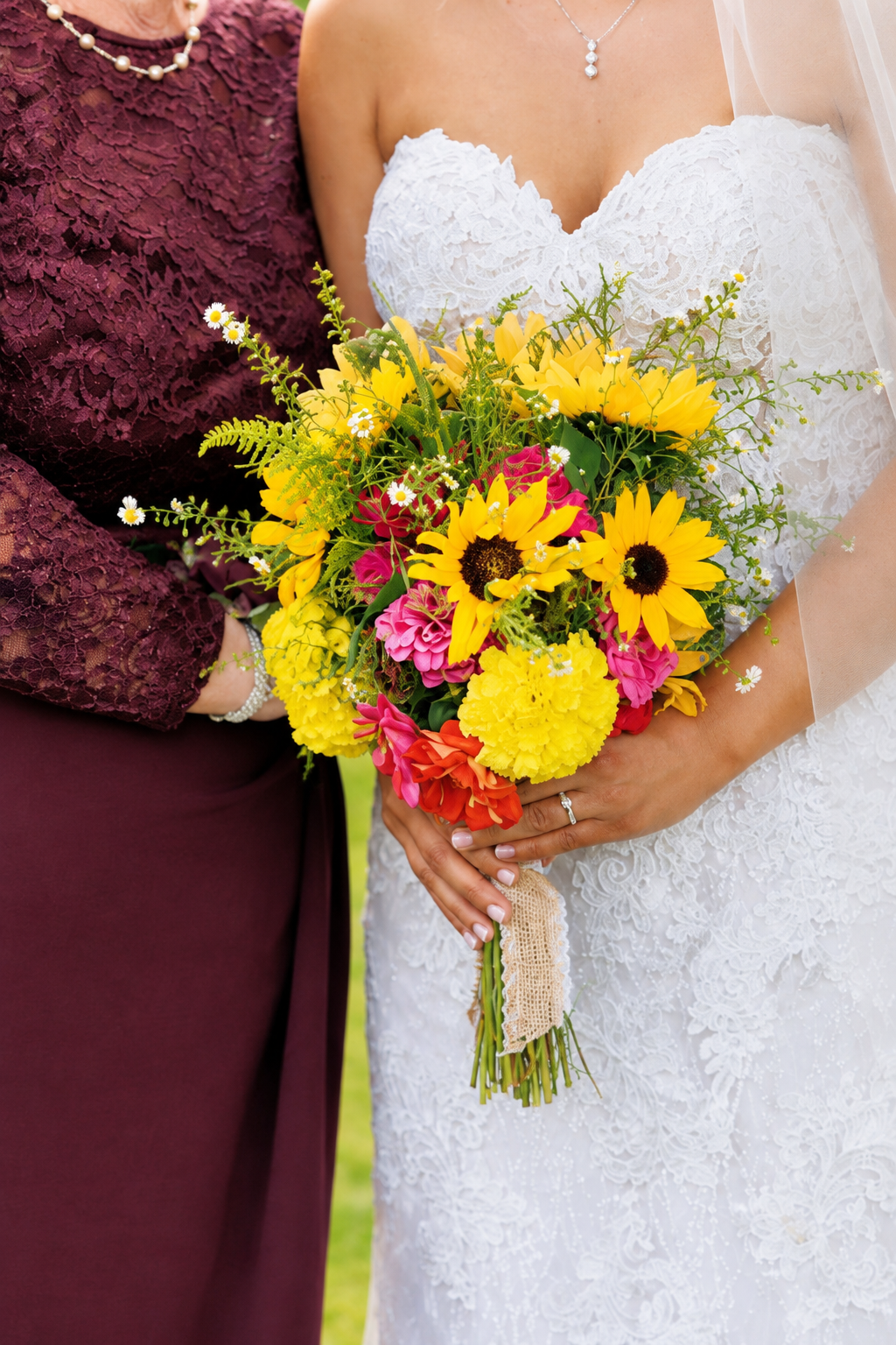 Close-up of a bride in a white lace wedding dress holding a colorful bouquet with yellow sunflowers, pink, yellow, and orange flowers, and green foliage, standing next to a woman in burgundy lace dress with pearl jewelry.