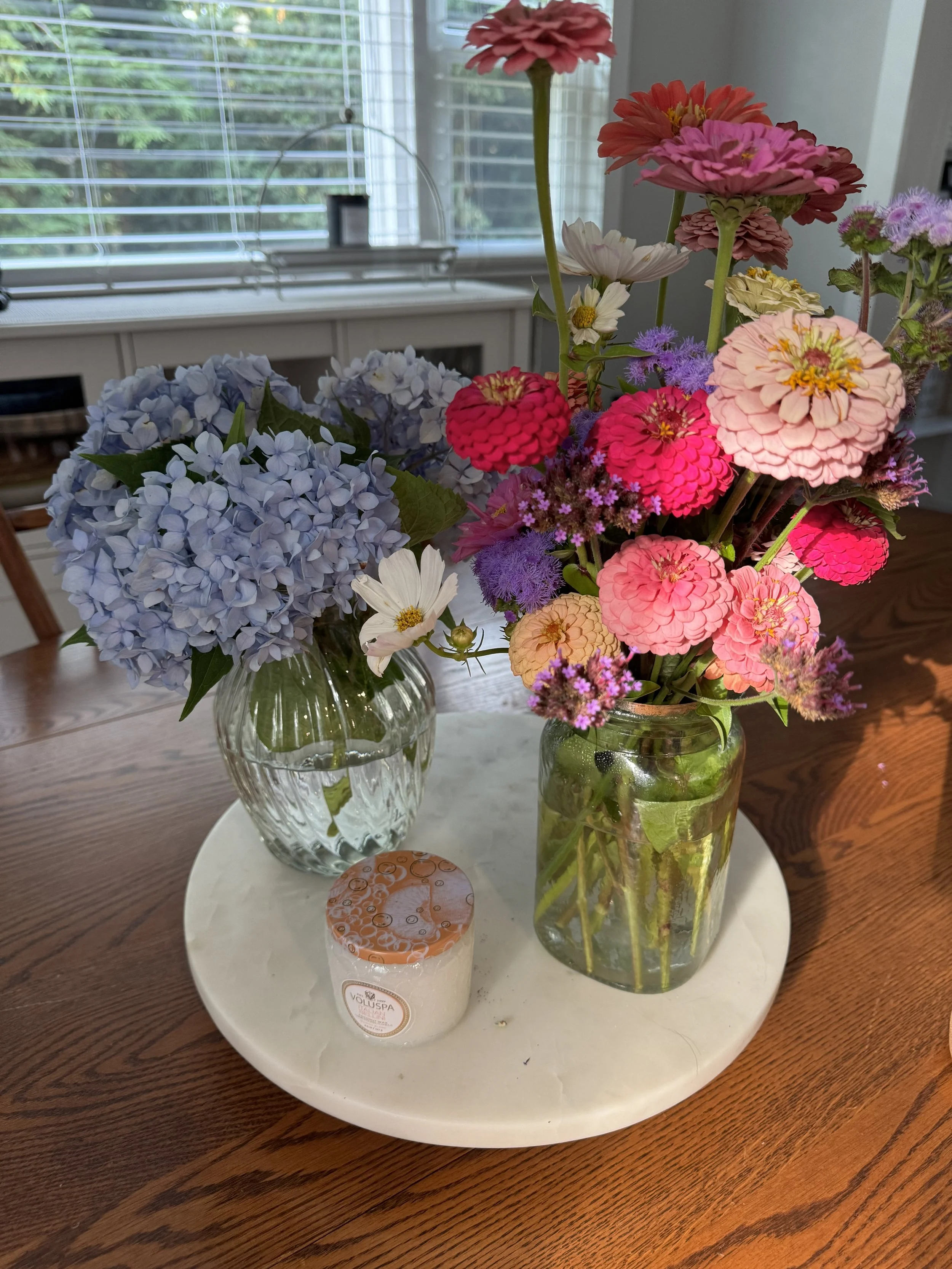 Two glass vases with colorful flowers, including hydrangeas and zinnias, placed on a white round tray on a wooden table near a window with blinds.
