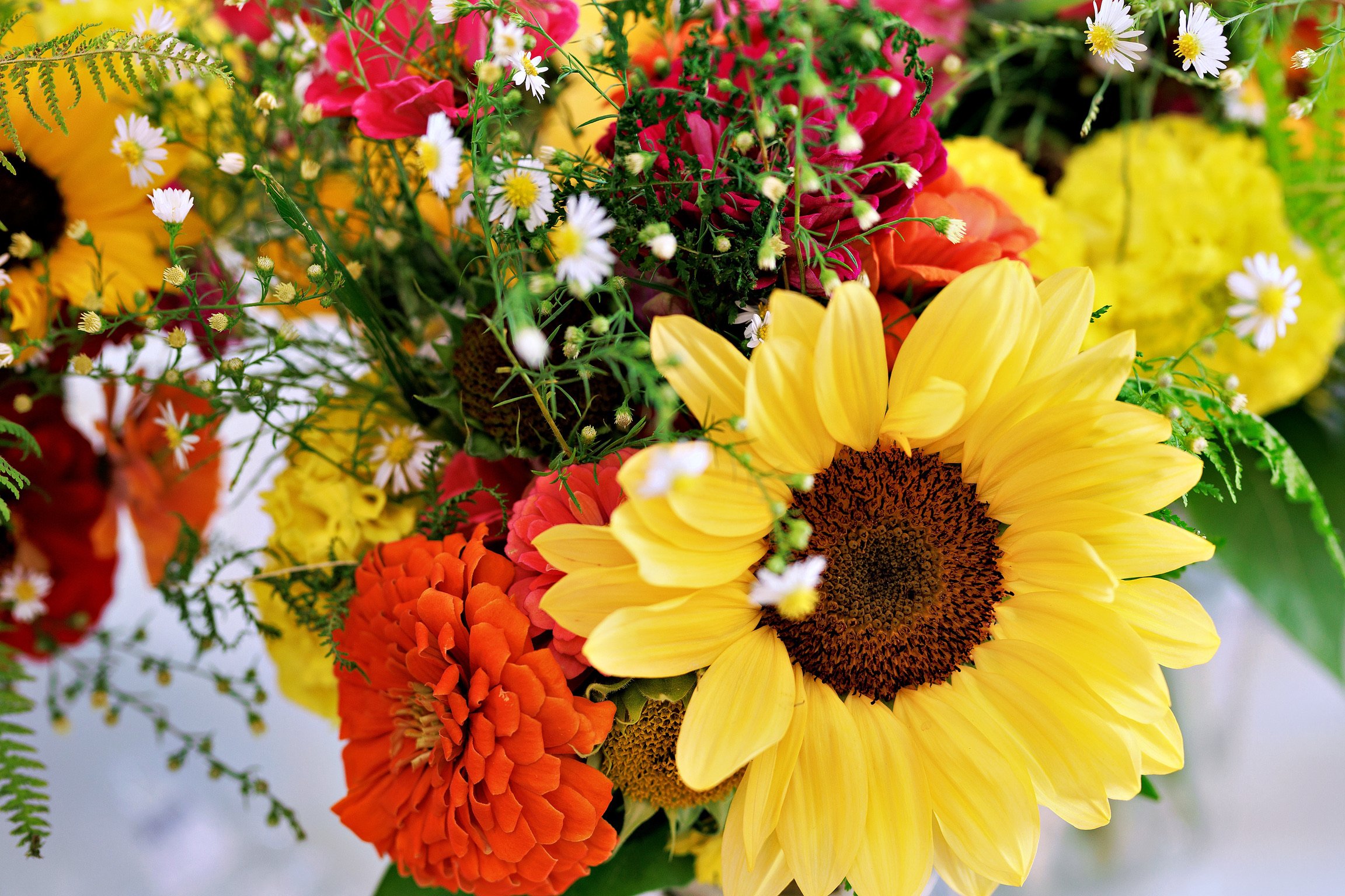 Close-up of a vibrant bouquet of flowers featuring a large yellow sunflower, small white daisies, orange dahlias, red and pink flowers, and various green foliage.