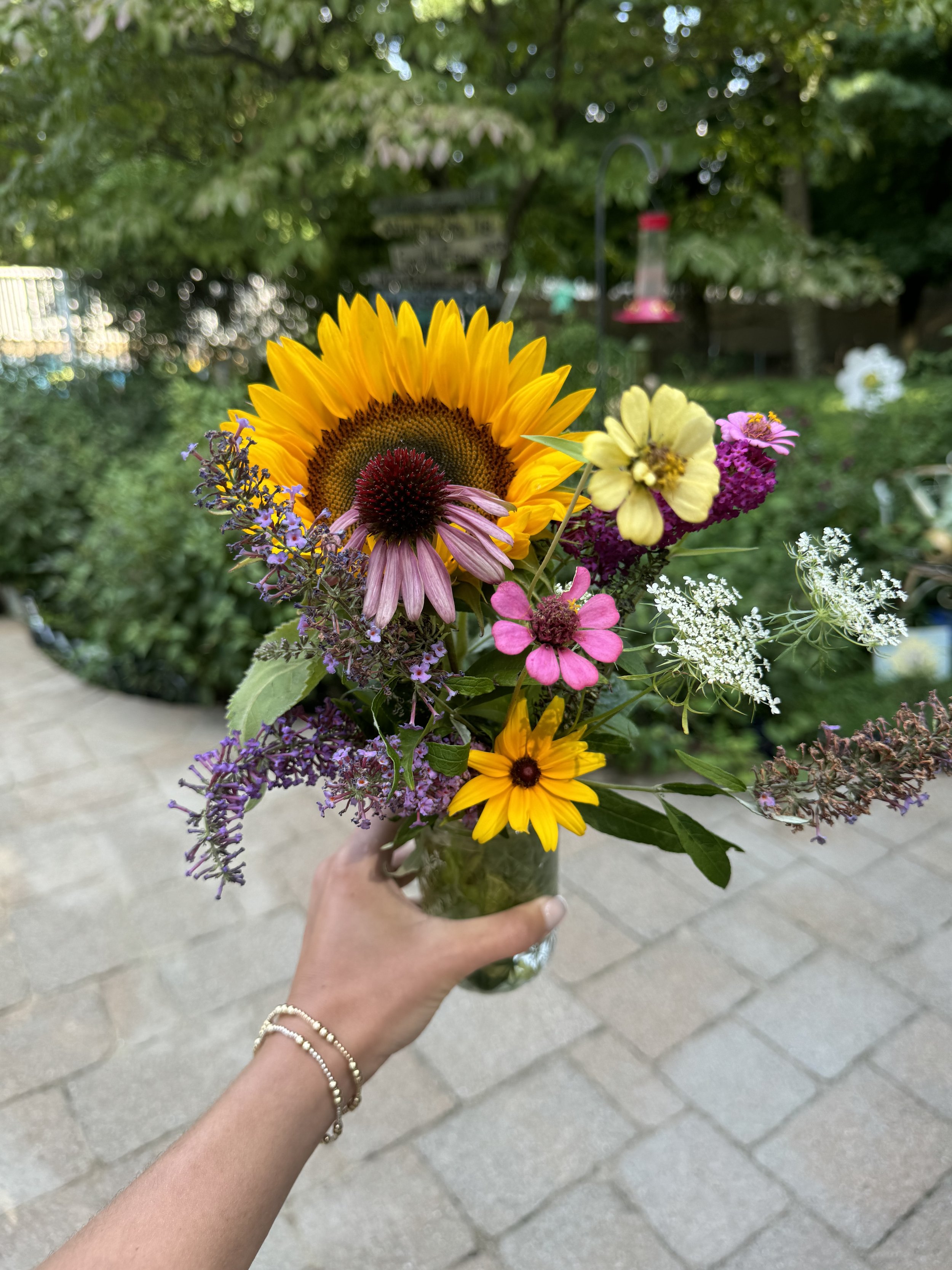 A person with a beige bracelet on their wrist holds a glass jar filled with a colorful bouquet of mixed flowers, including a large yellow sunflower, pink coneflowers, purple flowers, and small white flowers, outdoors with a garden and trees in the ba