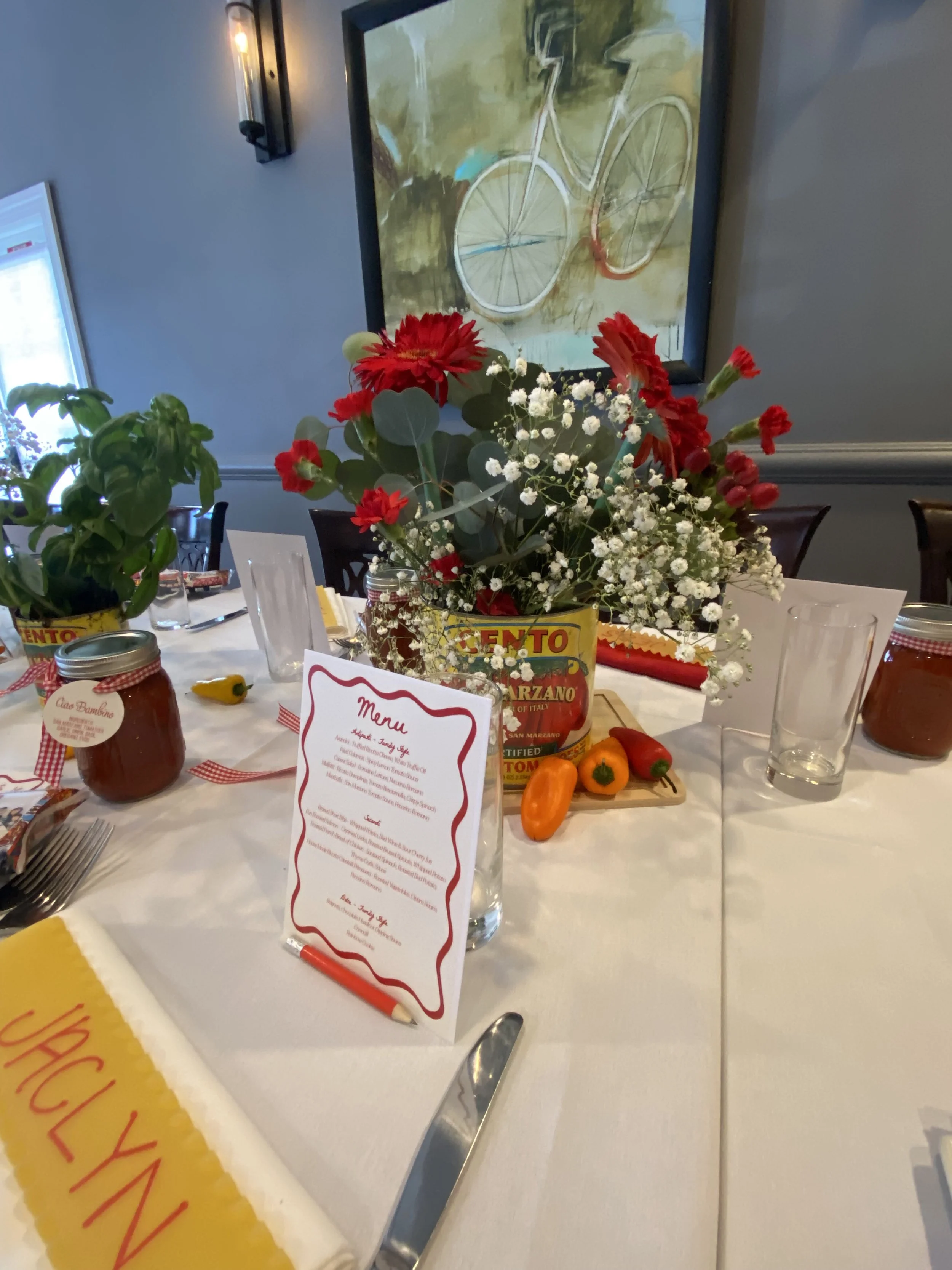 Table decorated with a floral centerpiece, jar of pasta sauce, a menu, and food items, set in a dining room with wall art and a window in the background.