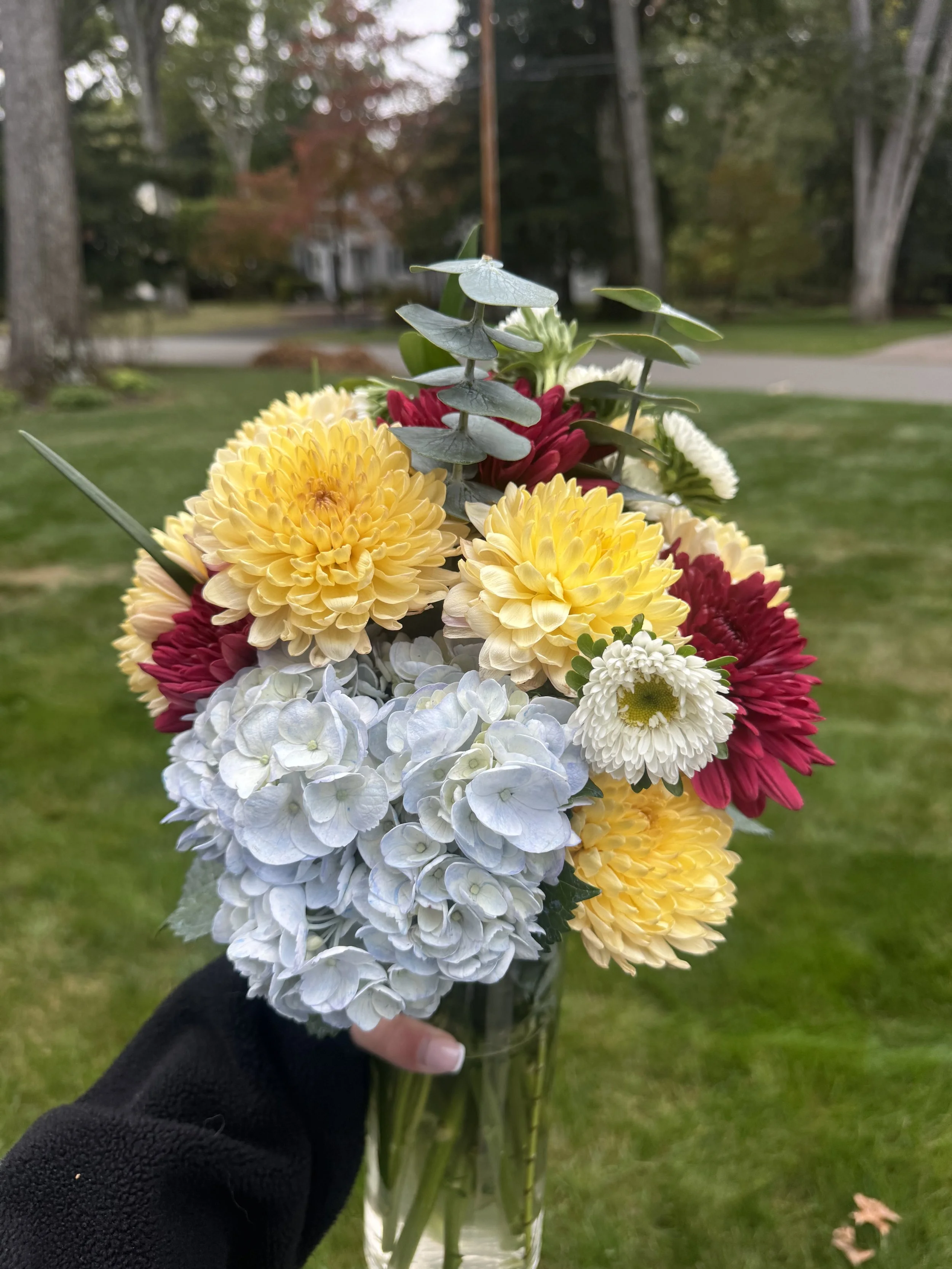 A person wearing a black coat holding a glass vase filled with a colorful bouquet of flowers, including white hydrangeas, yellow and red chrysanthemums, and red and white daisies, with a background of grass, trees, and a street.