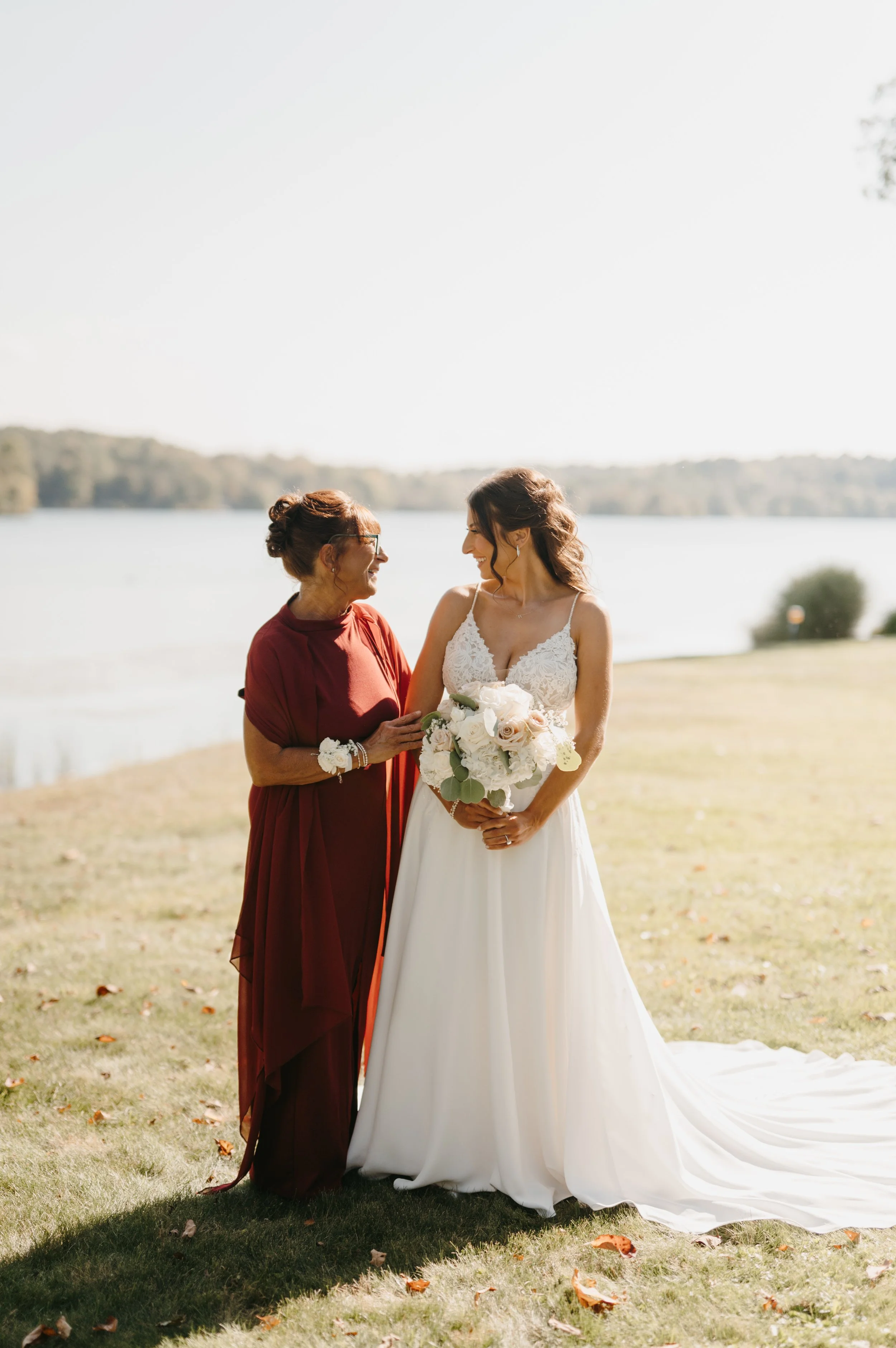 A bride and an older woman, possibly her mother, sharing a joyful moment outdoors during a wedding ceremony near a lake. The bride is in a white wedding dress holding a bouquet of white roses, and the older woman is in a dark red dress with a white corsage.