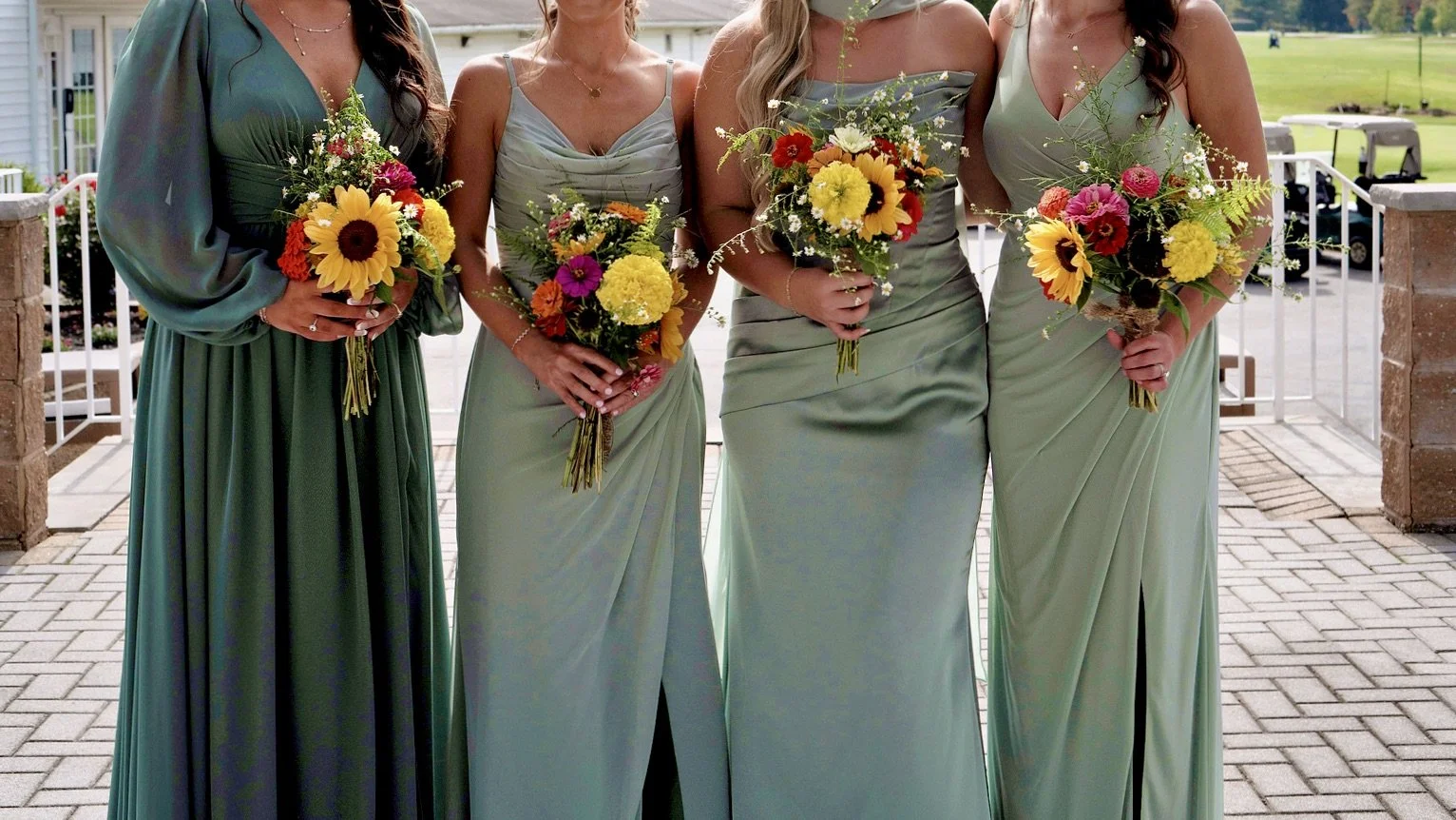 Four women in matching light green dresses holding colorful flower bouquets, standing outdoors on a paved area near a grassy field.