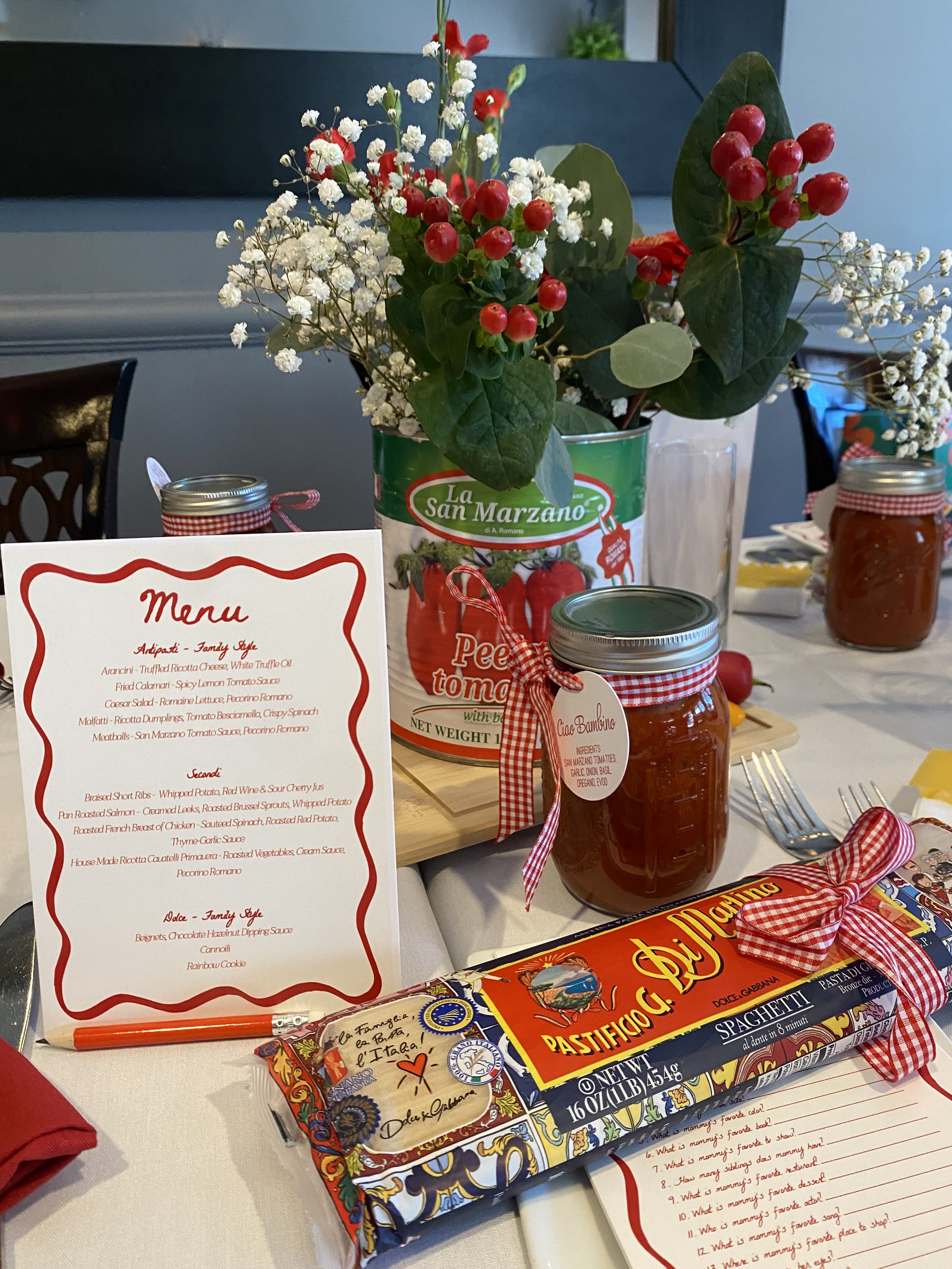 A tabletop display with a bouquet of white and red flowers in a can, jars of jam tied with red gingham ribbons, a menu, and a box of Italian pasta, with silverware and a red cloth on a white tablecloth.