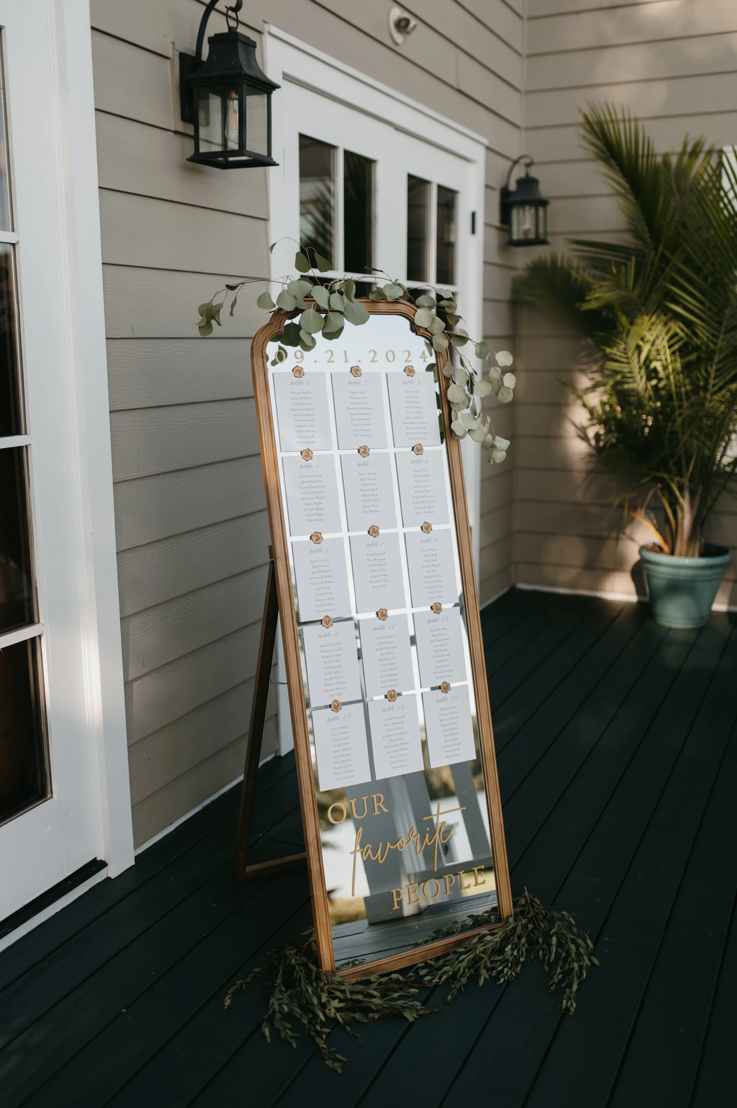 Wedding seating chart poster on an easel with decorative greenery, set outside on a porch with beige siding and black deck, next to a window and black lantern lights.