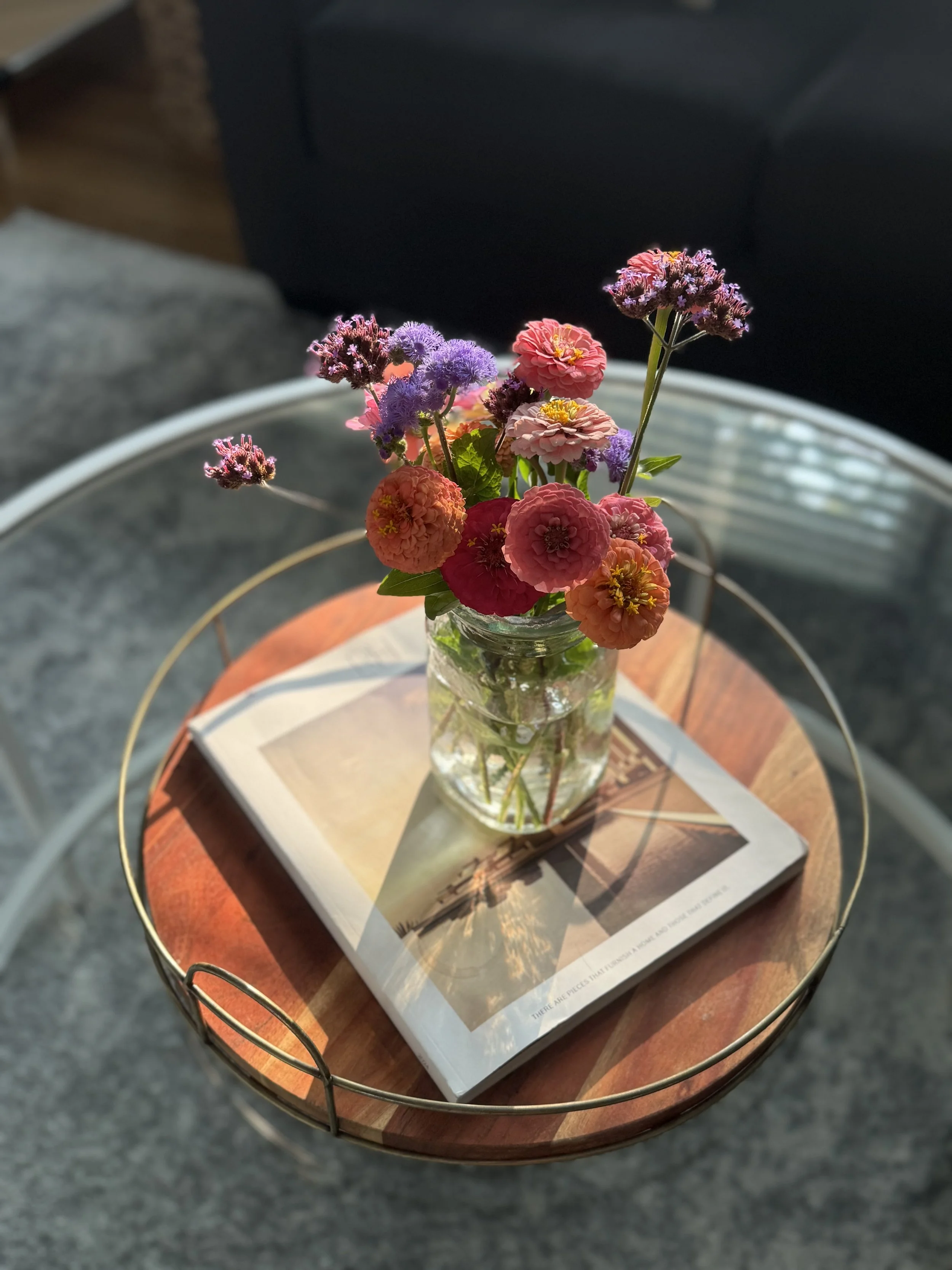 A glass jar of multicolored small flowers, including pink, purple, and orange blossoms, sitting on a magazine on a wooden table with a metal frame.