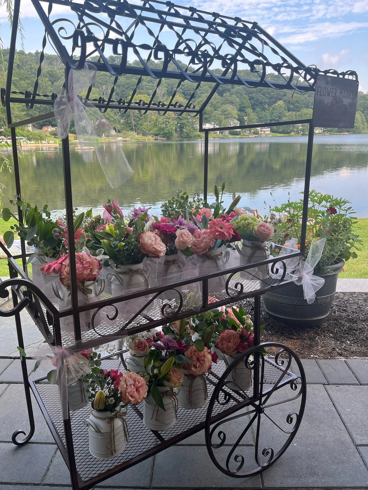 A decorative flower cart filled with pink and white flowers, set outdoors near a body of water with hills and trees in the background.