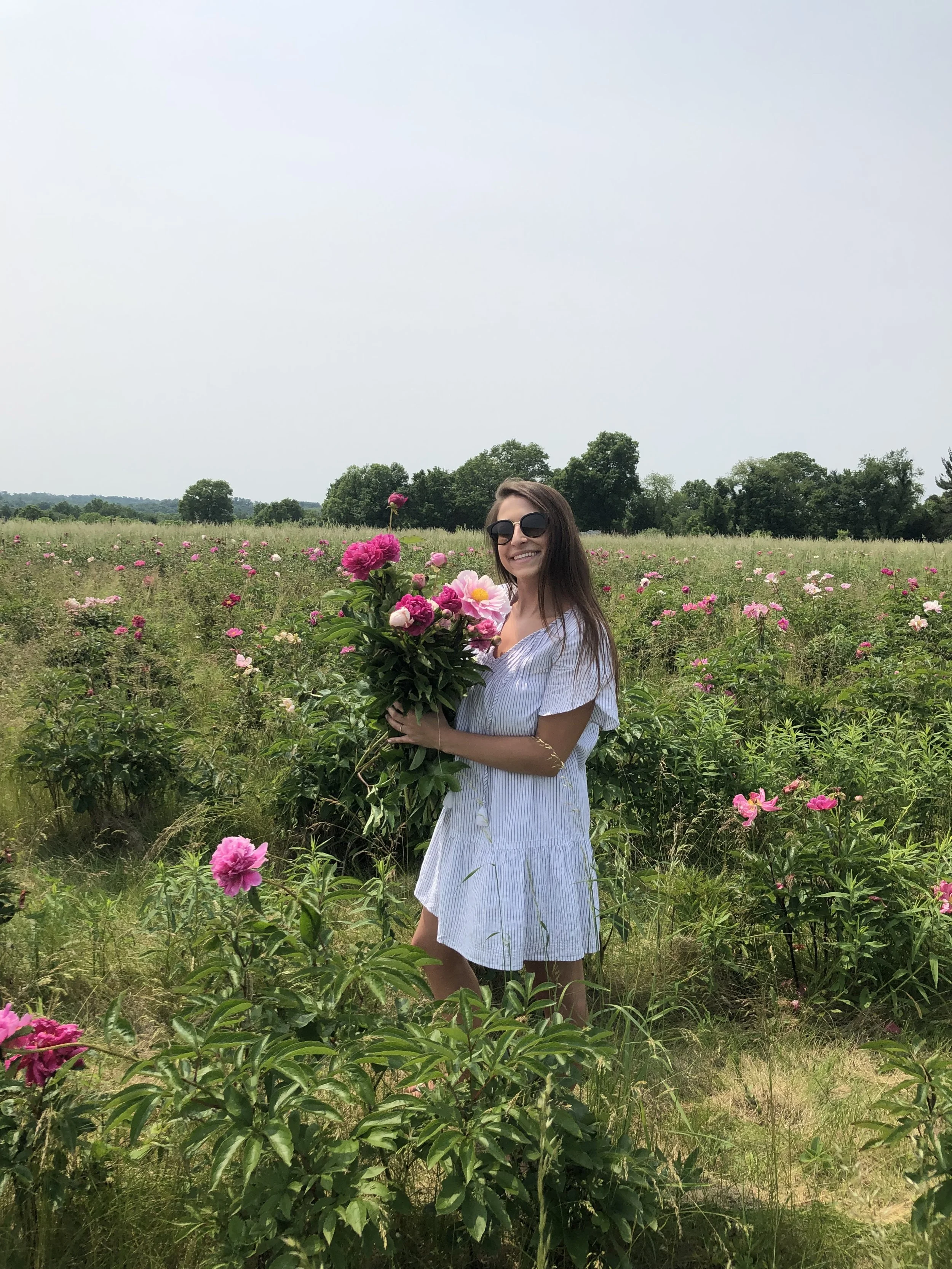 A woman in a white dress and sunglasses standing in a field of pink and white flowers, holding a bouquet of flowers, with trees in the background under a clear sky.