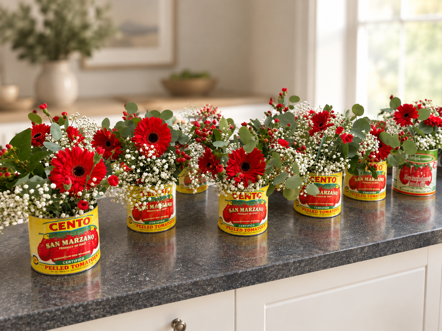 Six floral arrangements in canned tomatoes on a kitchen countertop with a white drawer, green leaves, red flowers, white baby's breath, in a bright kitchen with large windows.
