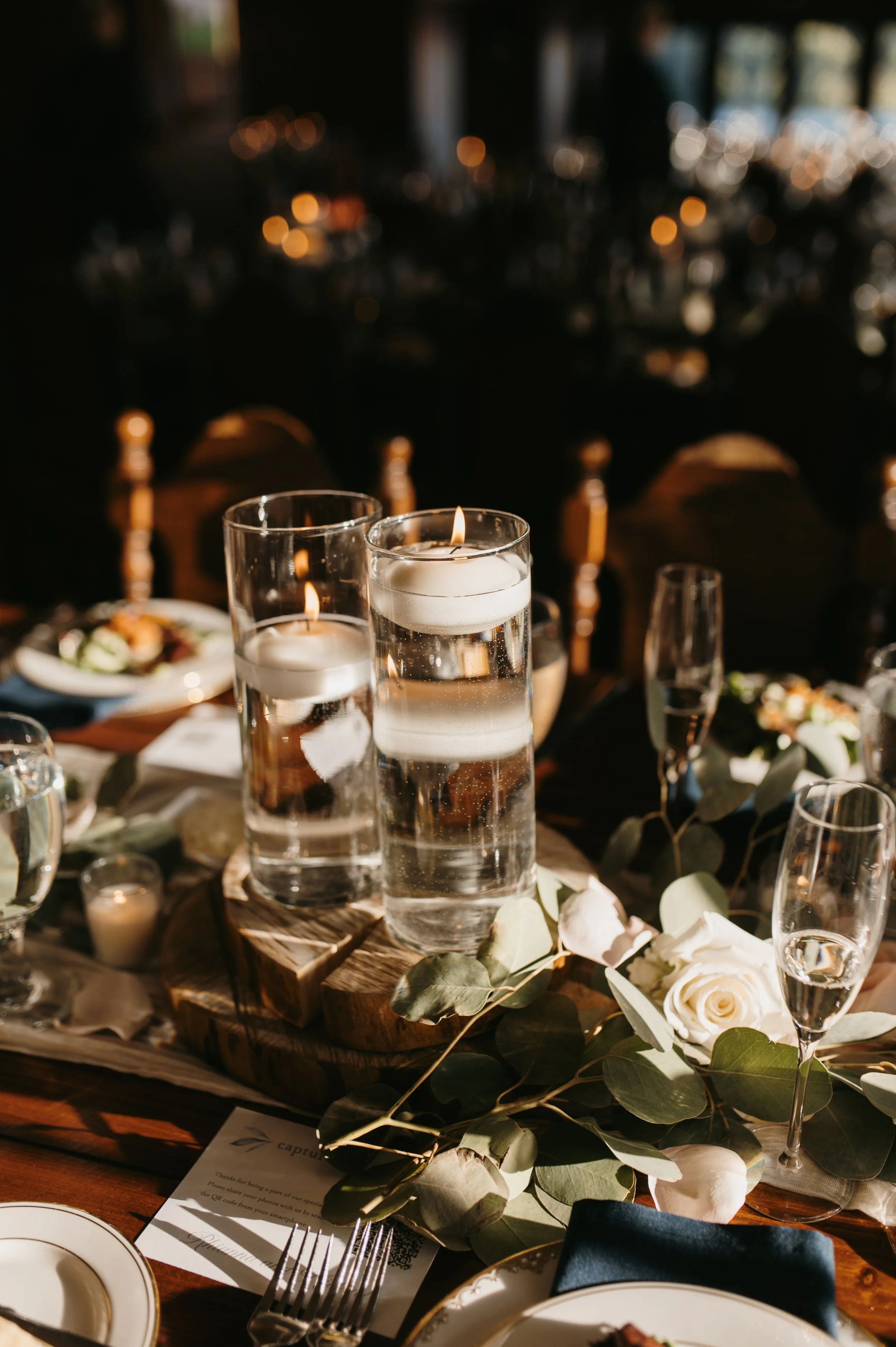 A table set for a formal event with candles, floral arrangements, white roses, and elegant dishes and glassware in a dimly lit room.